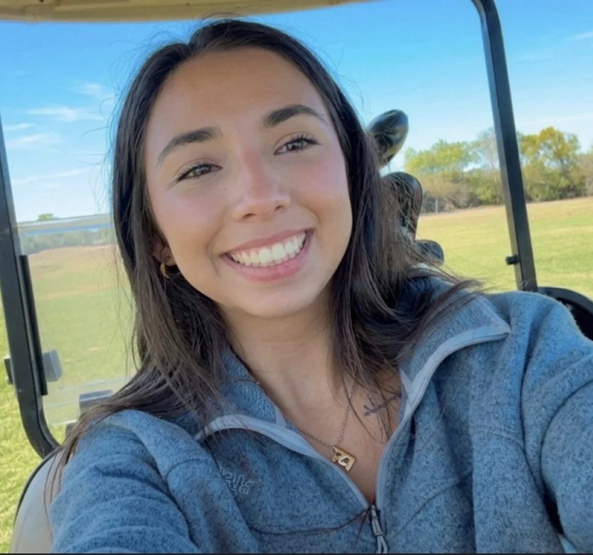 A young woman smiling for a selfie outdoors, sitting in a golf cart, with trees and blue sky in the background.