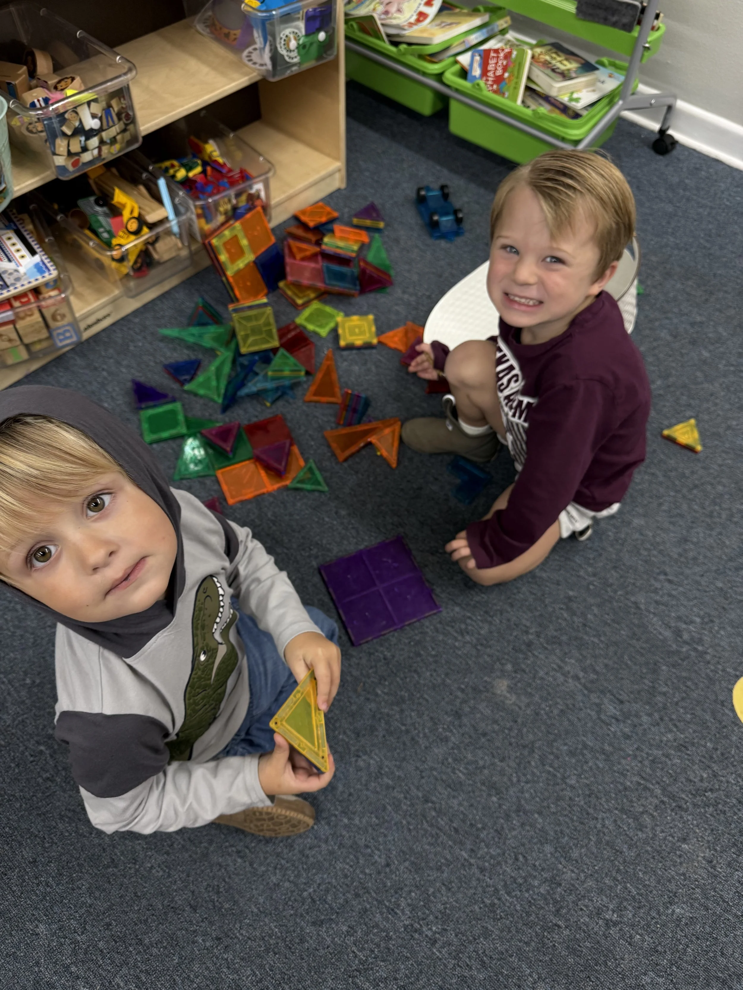 Two young boys playing with colorful magnetic building tiles on a carpeted floor in a classroom or playroom, with shelves of toys and books in the background.