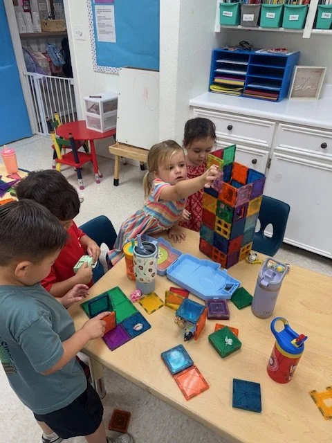 Four young children playing with colorful magnetic tiles on a table in a classroom.