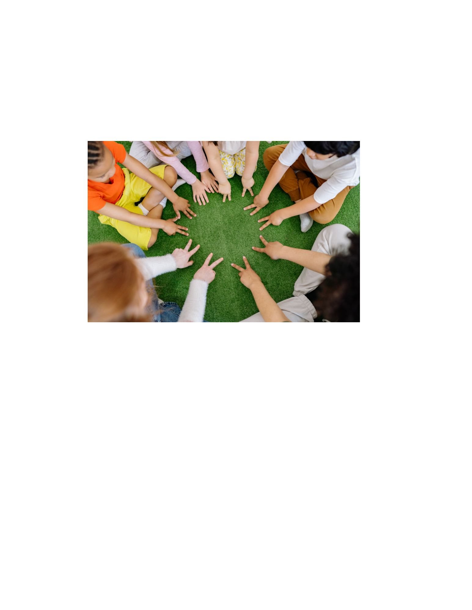 Children sitting in a circle on green carpet, pointing at each other with peace signs.