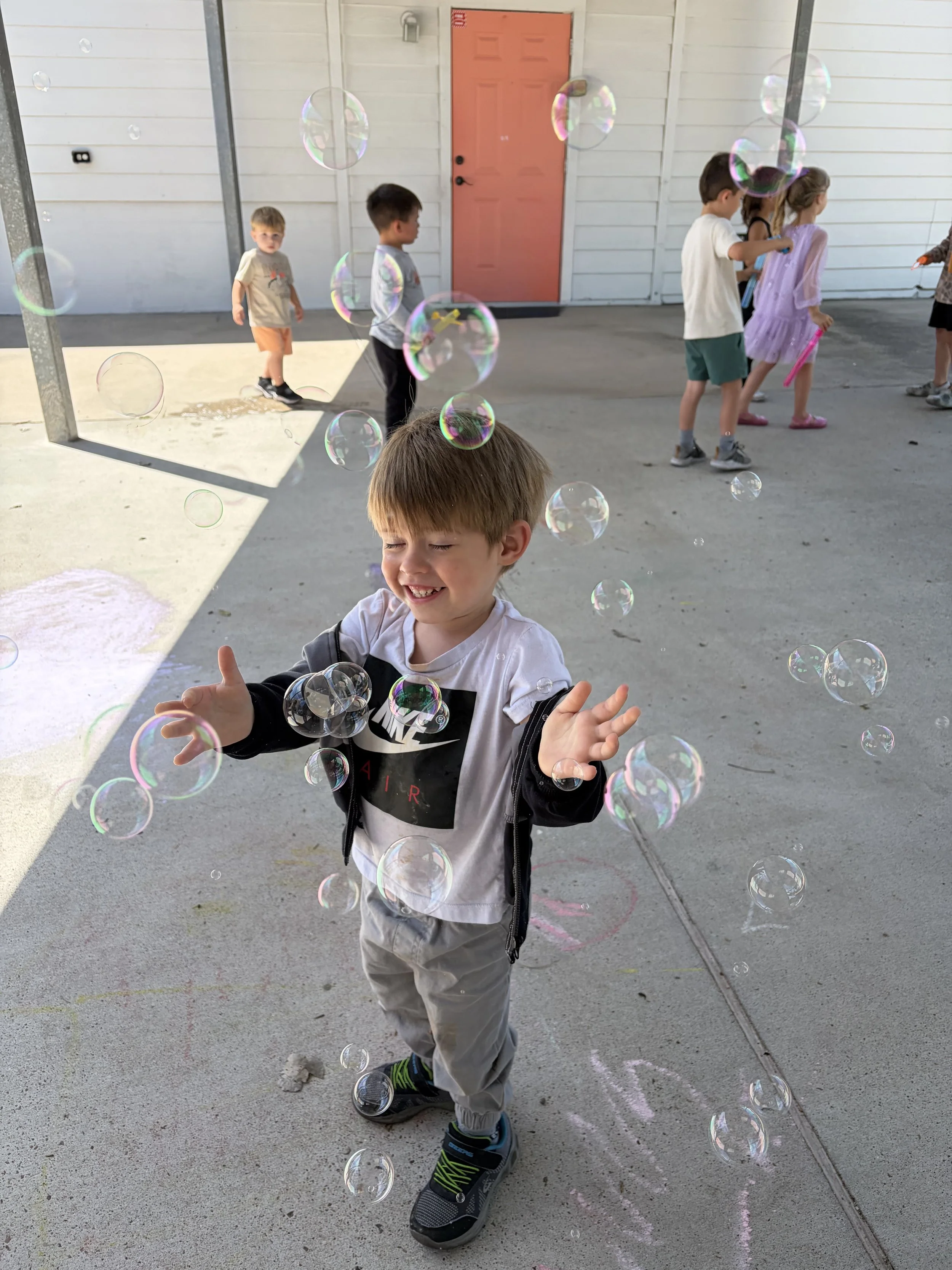 Young boy playing with bubbles outdoors, smiling with open arms, and laughing, surrounded by other children in the background.