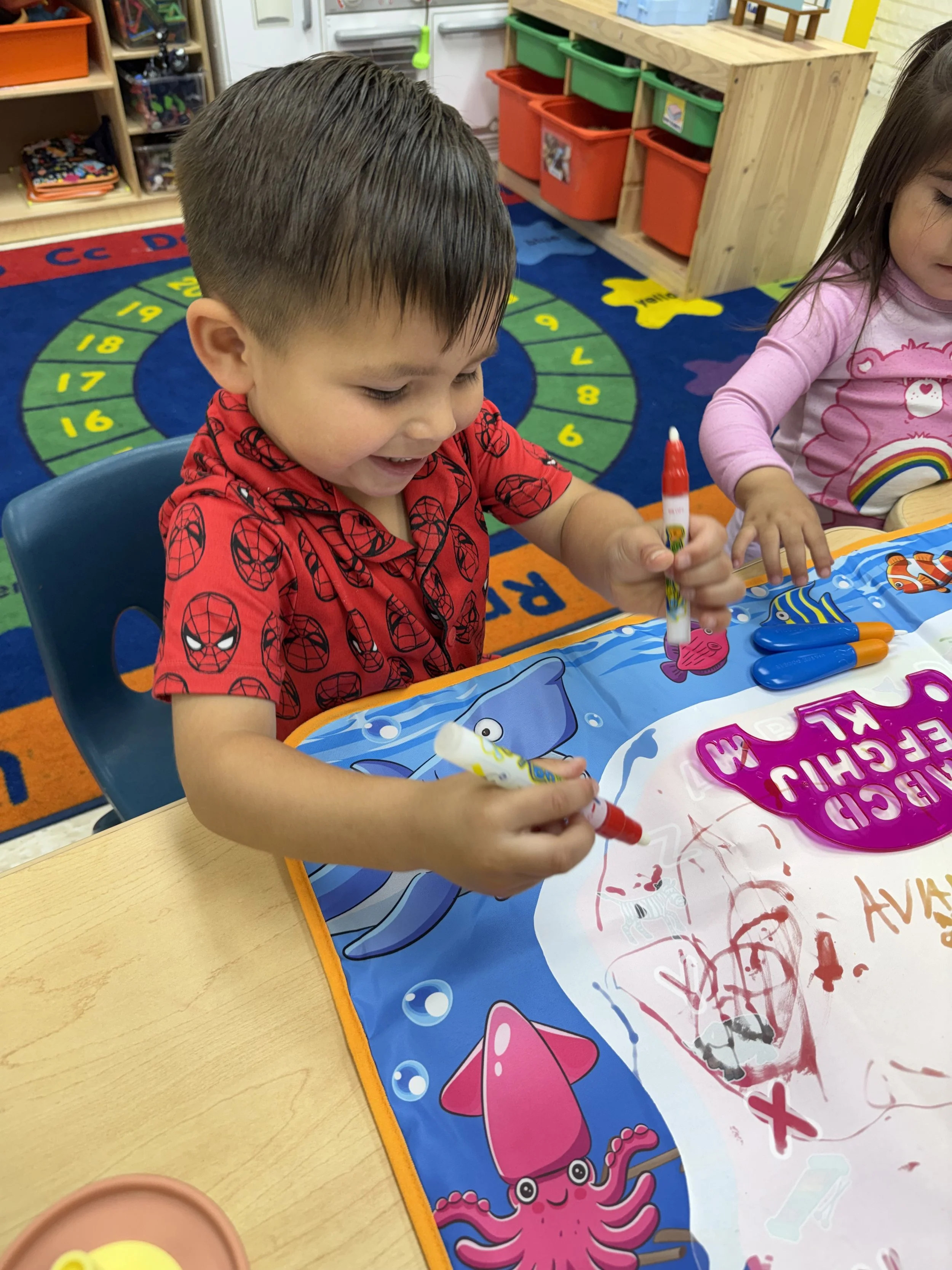 Two young children drawing on a paper with markers at a preschool table, with colorful educational decor in the background.