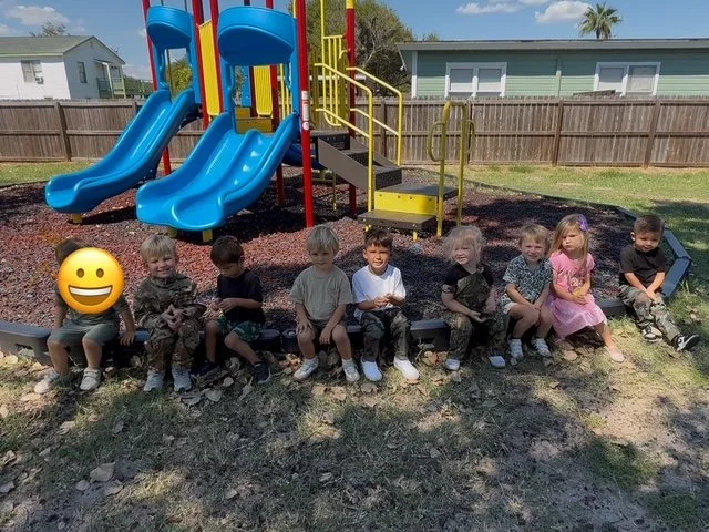 A group of young children sitting on a bench in front of a playground slide. The playground has two blue slides, yellow and red support structures, and a staircase. The children are outdoors on a sunny day, with a wooden fence and a house in the background.