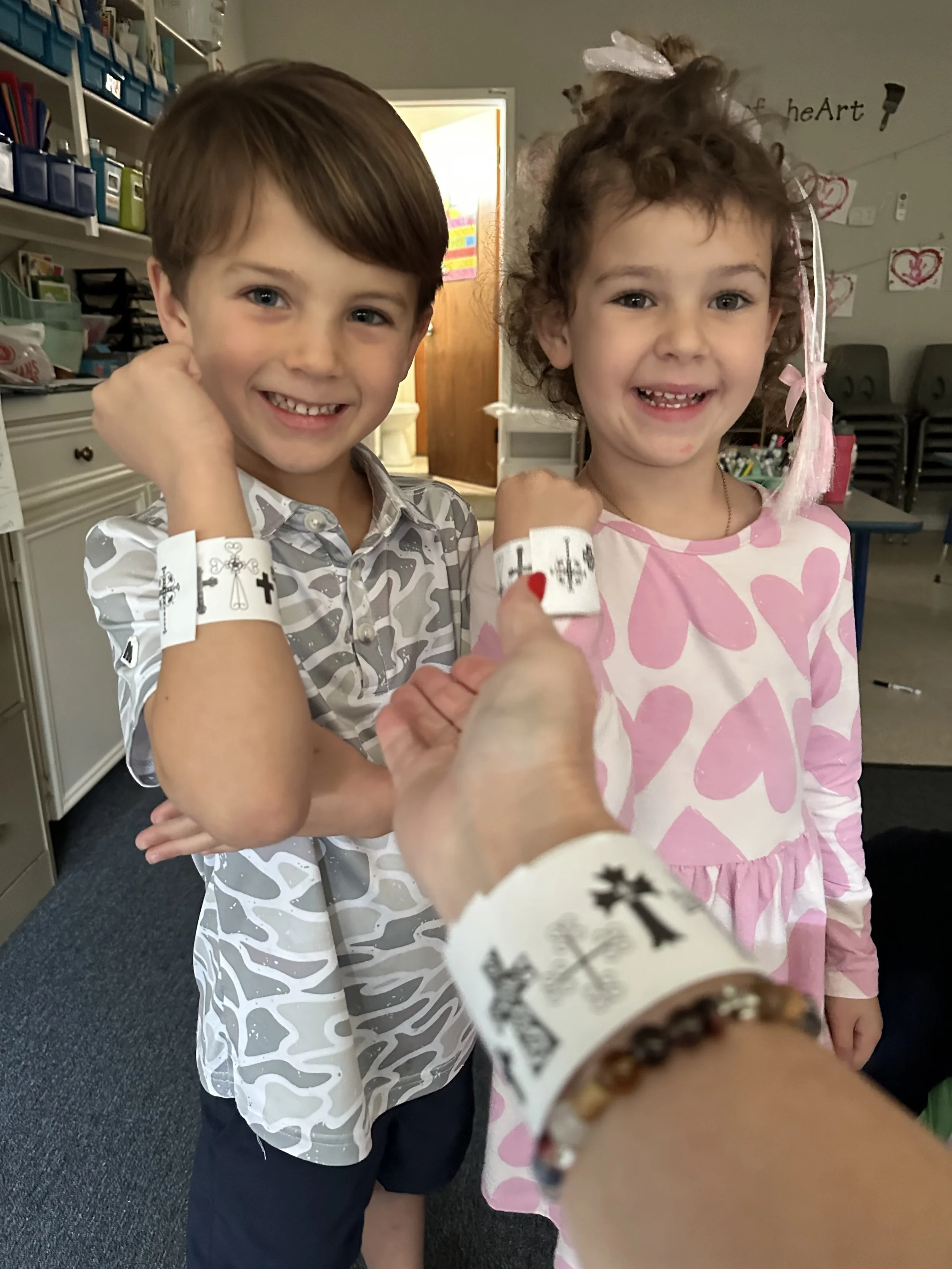 Two young children, a boy and a girl, smiling at camera, showing their hospital bracelets, in a classroom setting.