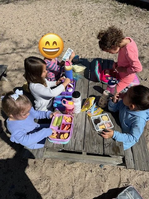 Four young girls sitting at a wooden picnic table outdoors, having a picnic with bento boxes and snacks, with one girl standing and all enjoying their time.