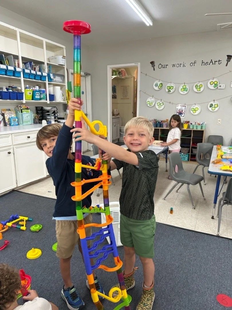 Two young boys in a classroom building a tall colorful tower with plastic building pieces, with a girl in the background at a table, and classroom shelves and wall decorations in view.
