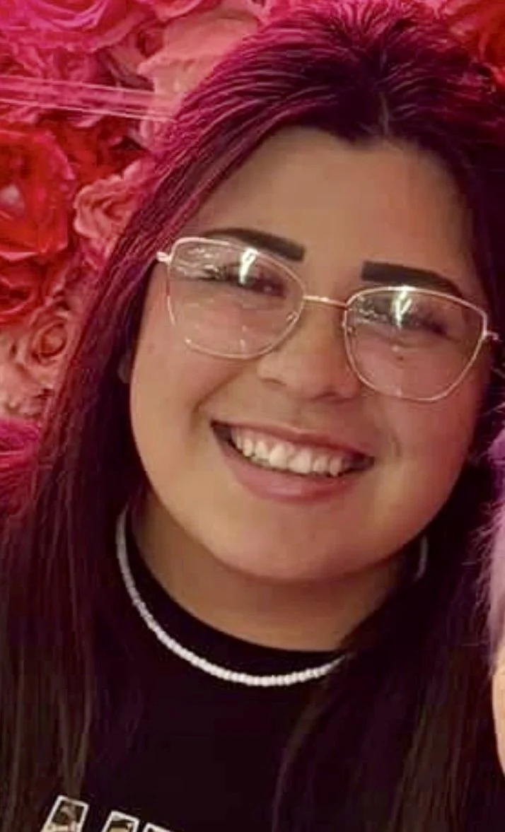 A young woman with long dark hair, glasses, dark eyebrows, and a bright smile, with pink roses in the background.