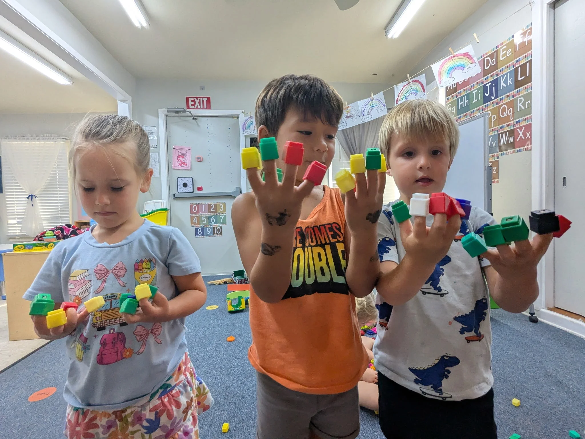 Three young children standing in a classroom, playing with colorful building blocks, with educational posters and alphabet charts on the walls.