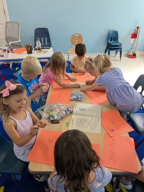 Children gathered around a table in a classroom, working on arts and crafts with orange paper, markers, and a scrapbook.