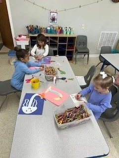 Three children sitting at a table engaged in arts and crafts activities in a classroom. They have colored paper, markers, and other craft supplies. One child is working with art supplies, while another is drawing or coloring.