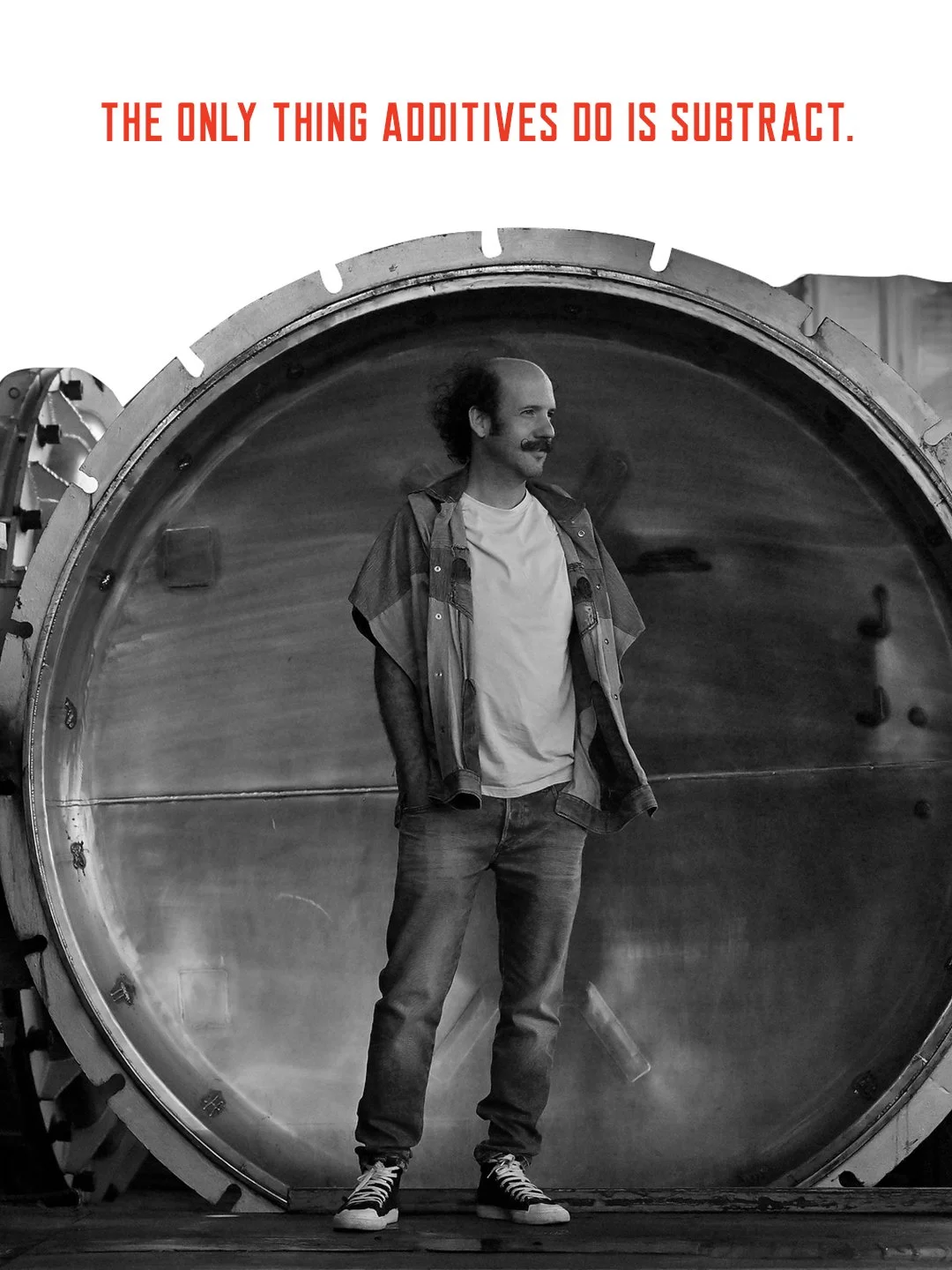 A man with a mustache and curly hair stands inside a large washing machine drum, wearing a casual white t-shirt, jeans, a plaid shirt, and sneakers, in a black and white photograph. Text above reads, 'The only thing additives do is subtract' in red font.