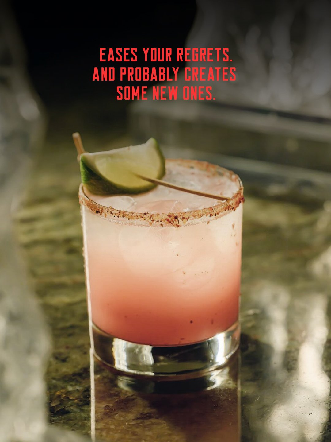 A pink cocktail in a short glass with a salted rim, garnished with a lime wedge, on a reflective surface with a blurred background. The text overlay reads, "Eases your regrets. And probably creates some new ones."