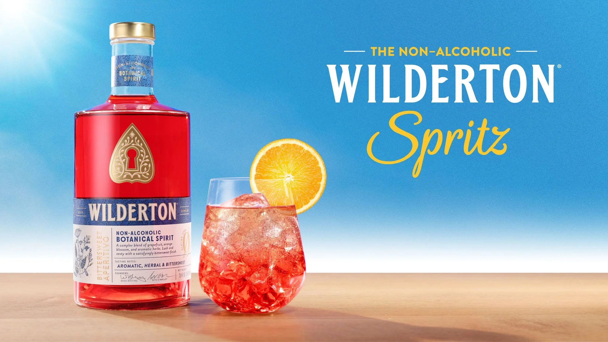Bottle of Wilderton Spritz non-alcoholic botanical spirit with a glass of pink drink garnished with an orange slice, against a blue sky background with the text 'The Non-Alcoholic Wilderton Spritz'.