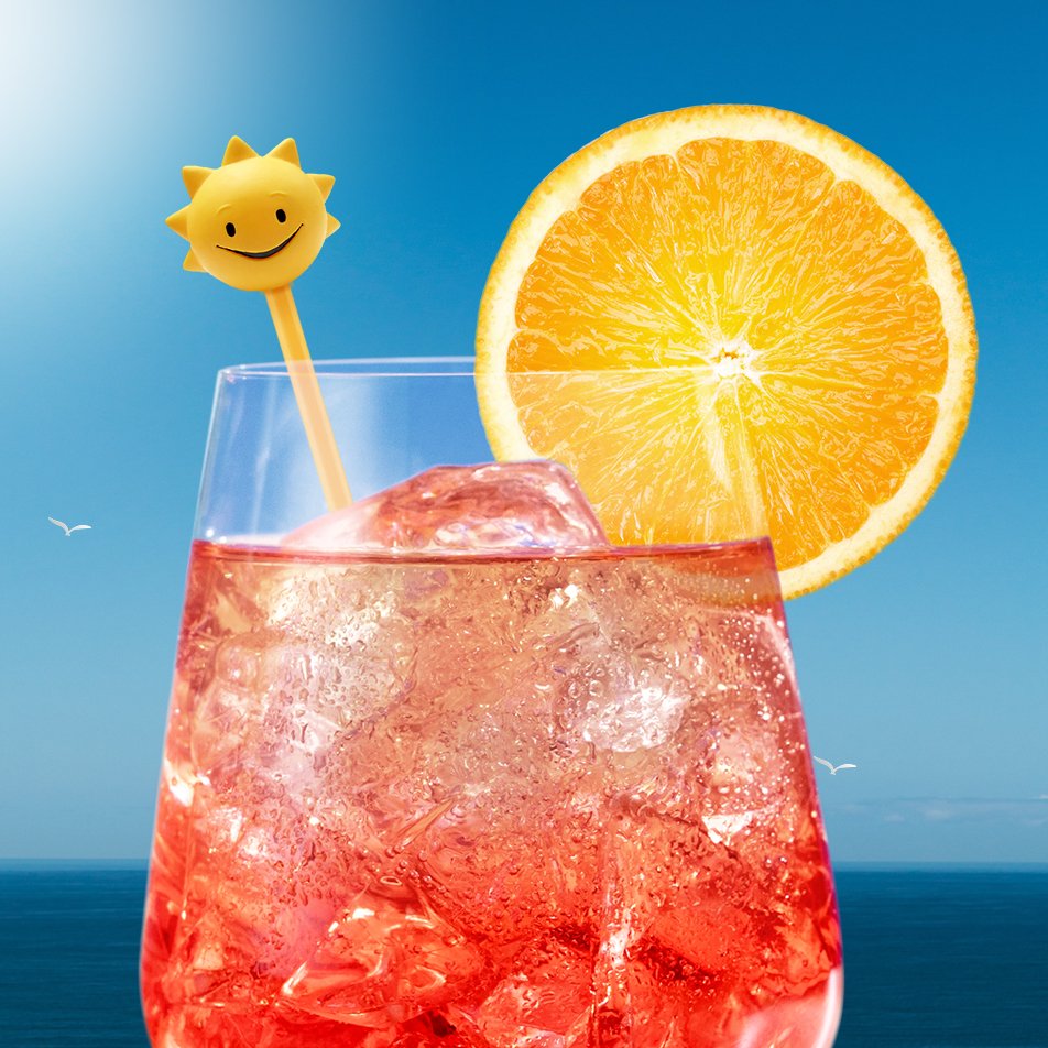 A pink cocktail with ice cubes in a clear glass garnished with a slice of orange and a smiling sun-shaped drink stirrer, set against a blue sky background with the ocean visible at the bottom.