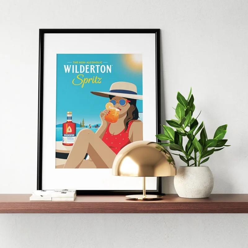A framed poster of a woman in a red swimsuit and large sun hat at the pool, drinking a cocktail, with a bottle of Wilderton Spritz in the background, placed on a wooden shelf alongside a gold dome lamp and a potted green plant.