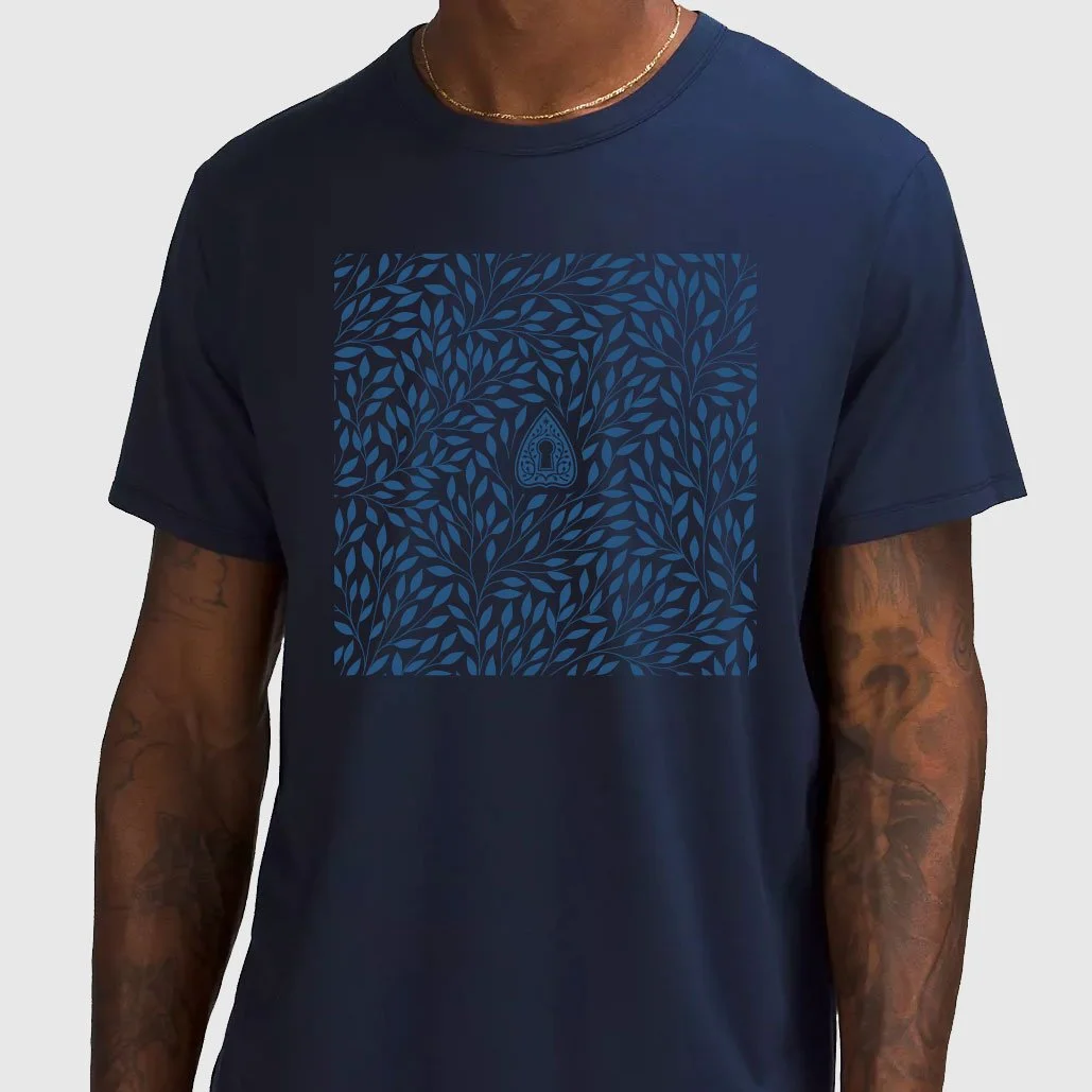 A man wearing a navy blue T-shirt with a dark blue leafy pattern and a small symbol in the center, and a gold necklace.