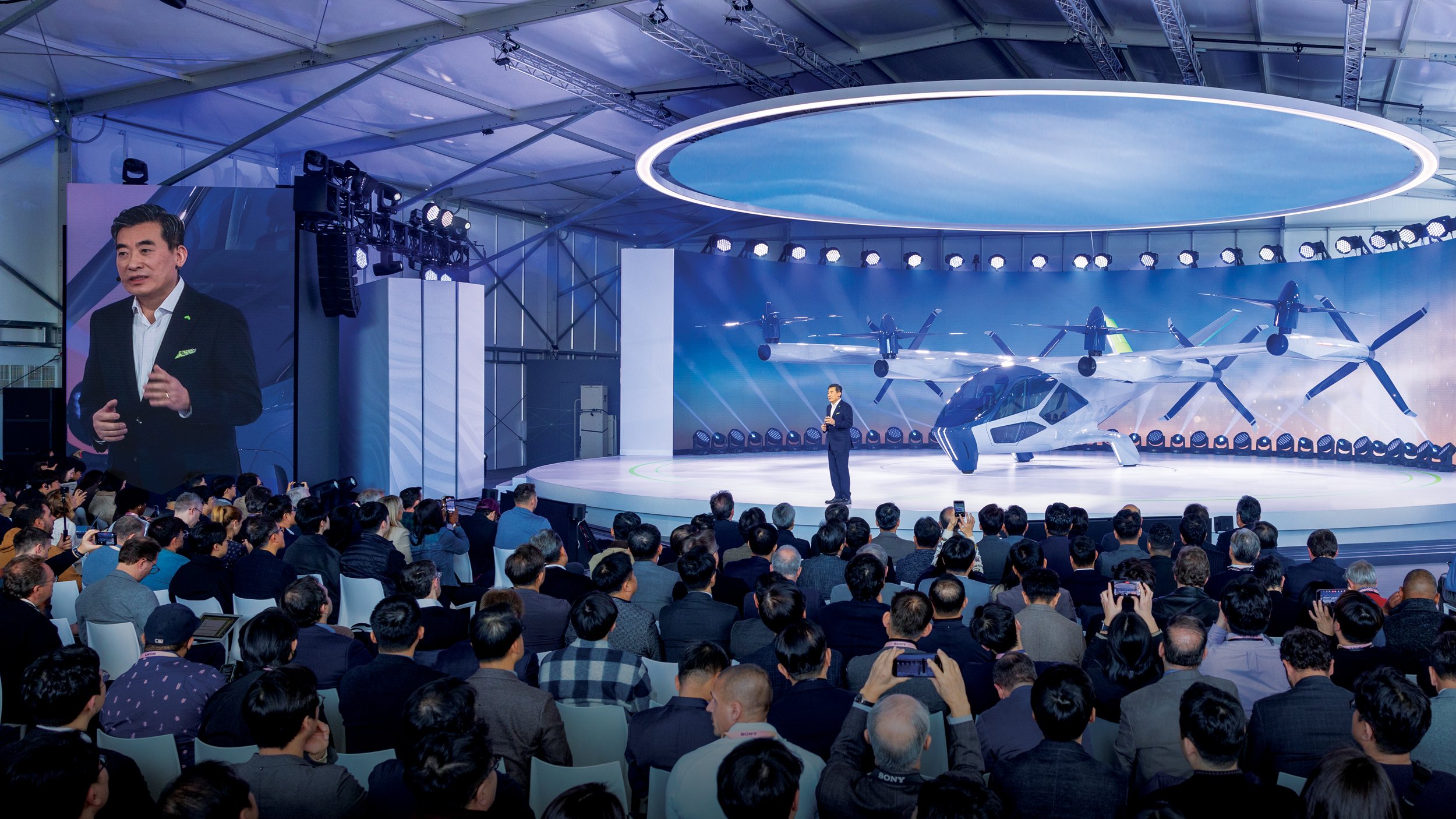 A man in a suit giving a presentation at a conference with a large audience. Behind him on stage, there are futuristic aircraft models and a digital display with clouds, under a circular light fixture on the ceiling.