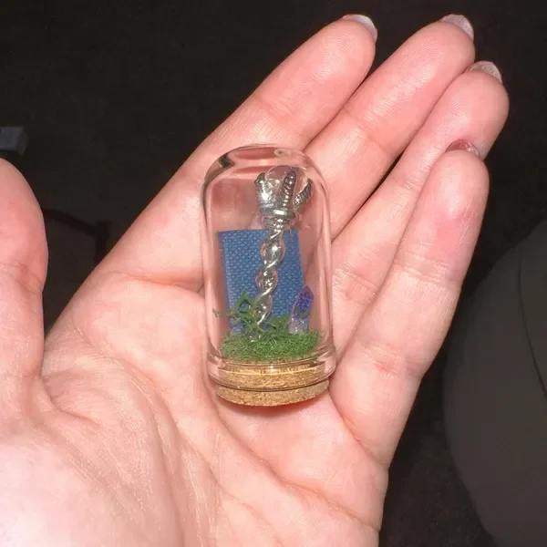 A small glass container with a cork base holding a miniature silver twisted rod, green decorative moss, and a blue background piece.