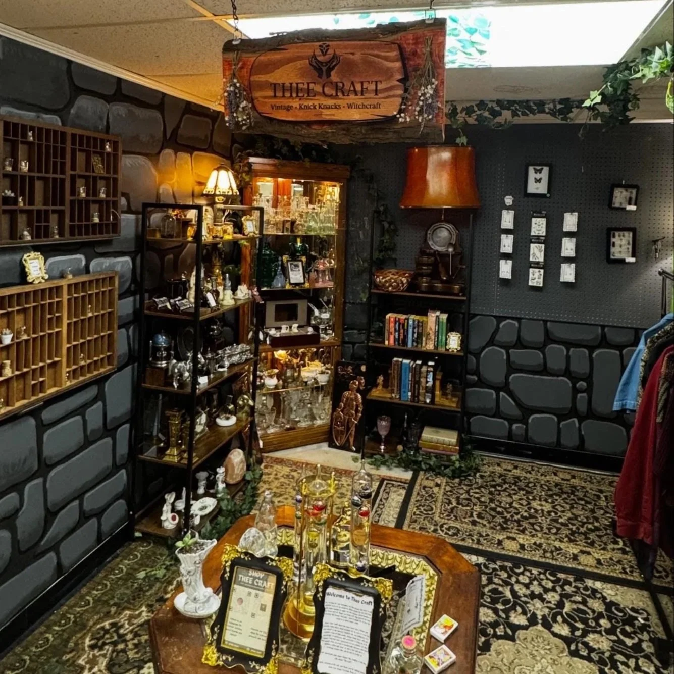 Interior of a vintage thrift and craft store with dark stone walls, wooden display shelves, glassware, books, and decorative items on display. A sign hanging from the ceiling reads 'THEE CRAFT.' Baroque-style patterned carpet covers the floor, and there are decorative plants and framed art on the walls.