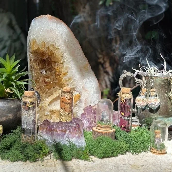 A decorative display featuring a large yellowish geode, several small glass vials with cork stoppers containing various small objects, a cluster of purple and clear crystals, green moss, and a potted succulent plant.