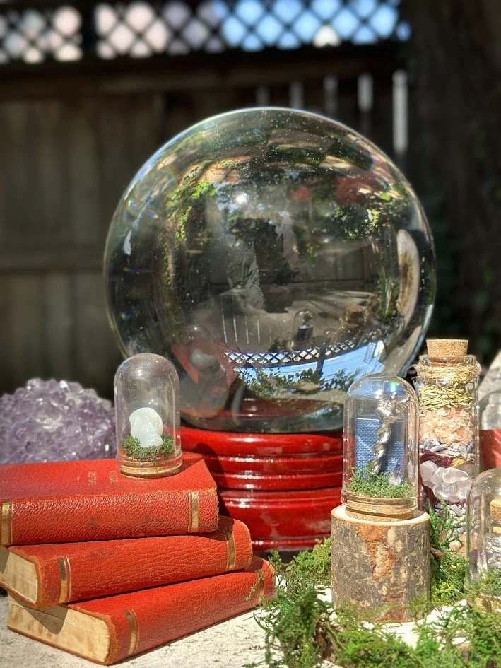 Decorative display with a large glass sphere, several glass jars with miniature plants, books with red covers, and crystals outdoors on a table.