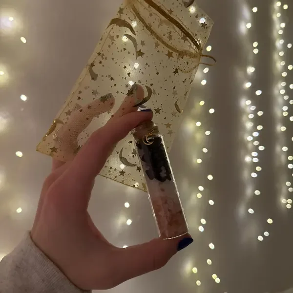 A person's hand holding a clear tube with a dark substance inside, partially wrapped in festive gold wrapping paper with star and moon patterns, in front of a background of blurred string lights.