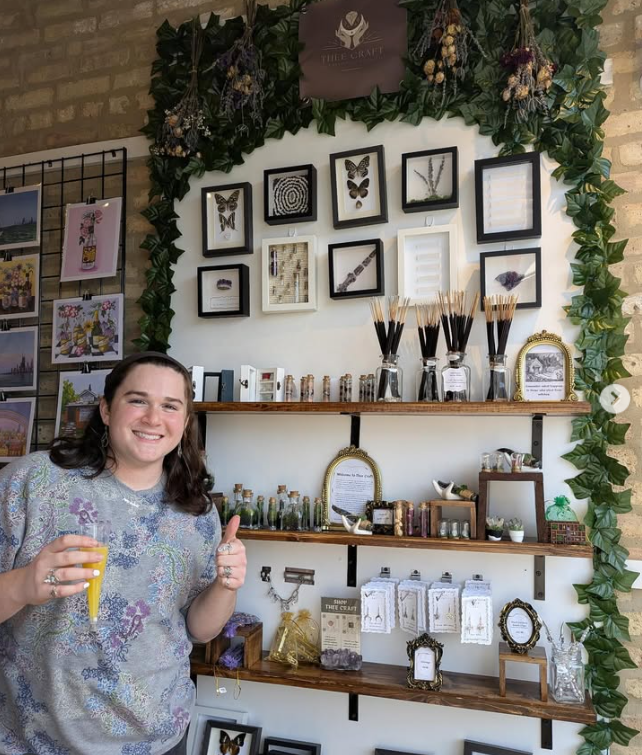 A woman smiling and giving a thumbs-up in front of a display wall with framed art, small bottles, and decorative items at an indoor market or shop.