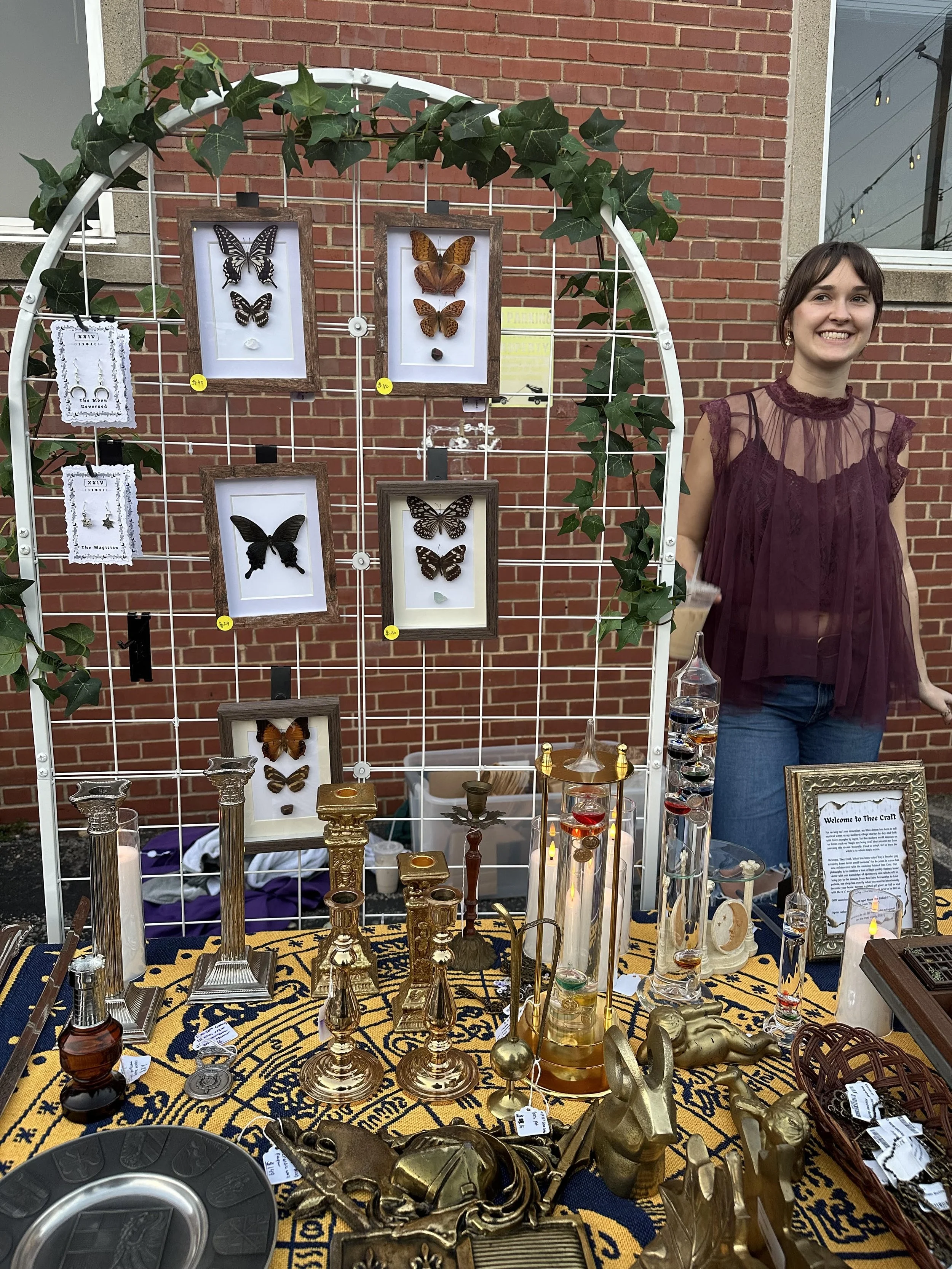 An outdoor craft sale table displaying framed butterfly and insect displays, brass and gold decorative items, glass vials, and a framed welcome sign, with a smiling young woman standing beside it against a brick wall.