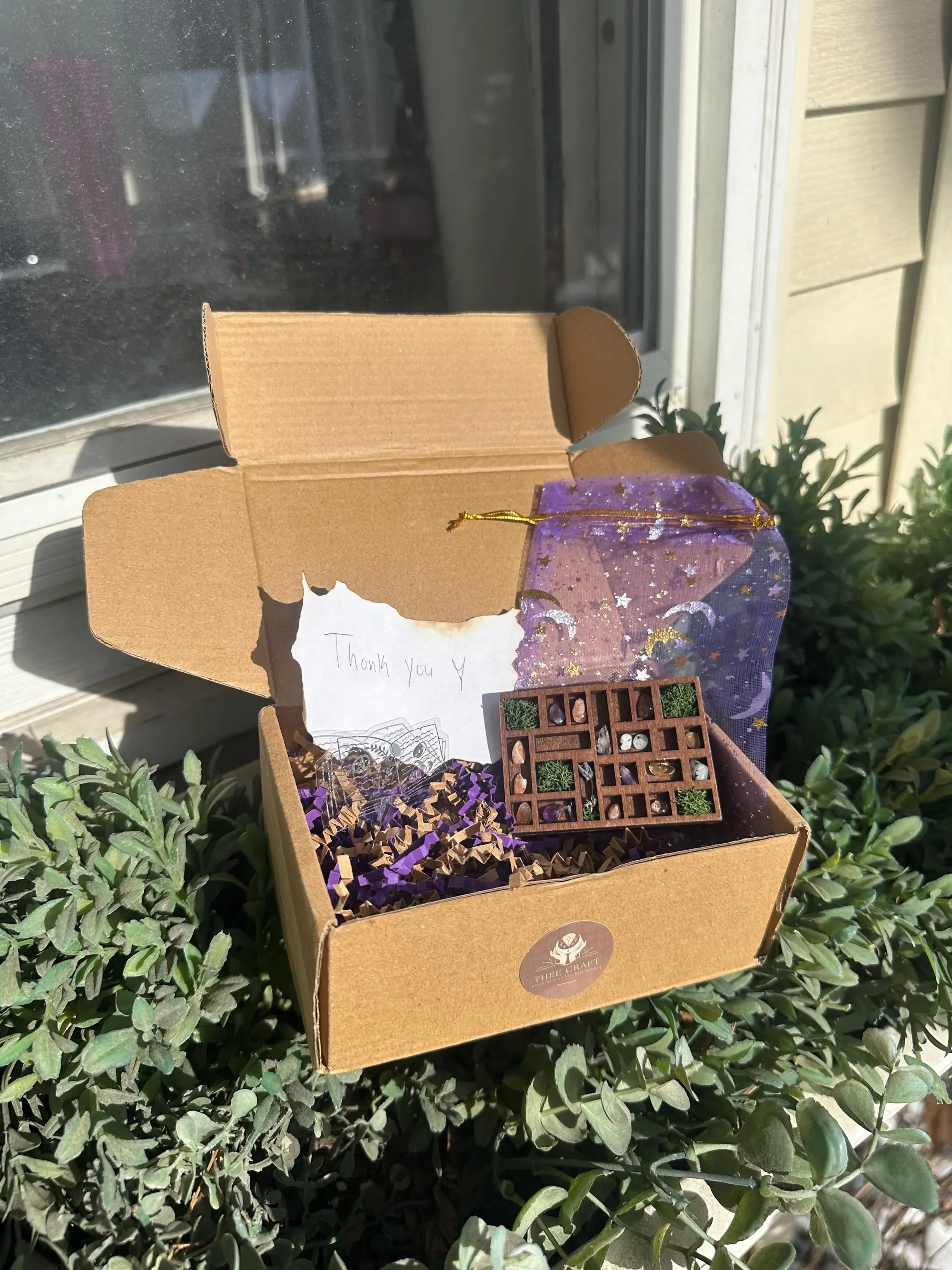 A cardboard box containing a thank you note, some purple shredded paper, small plants, and a miniature wooden maze puzzle with various small items inside, set on green foliage outside near a house window.