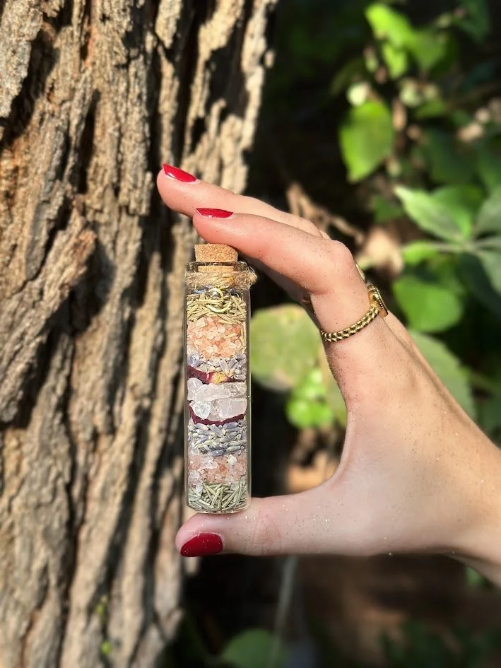 Hand with red nail polish holding a small glass jar filled with layered colorful herbs and salts. The background shows a tree trunk and green leaves.