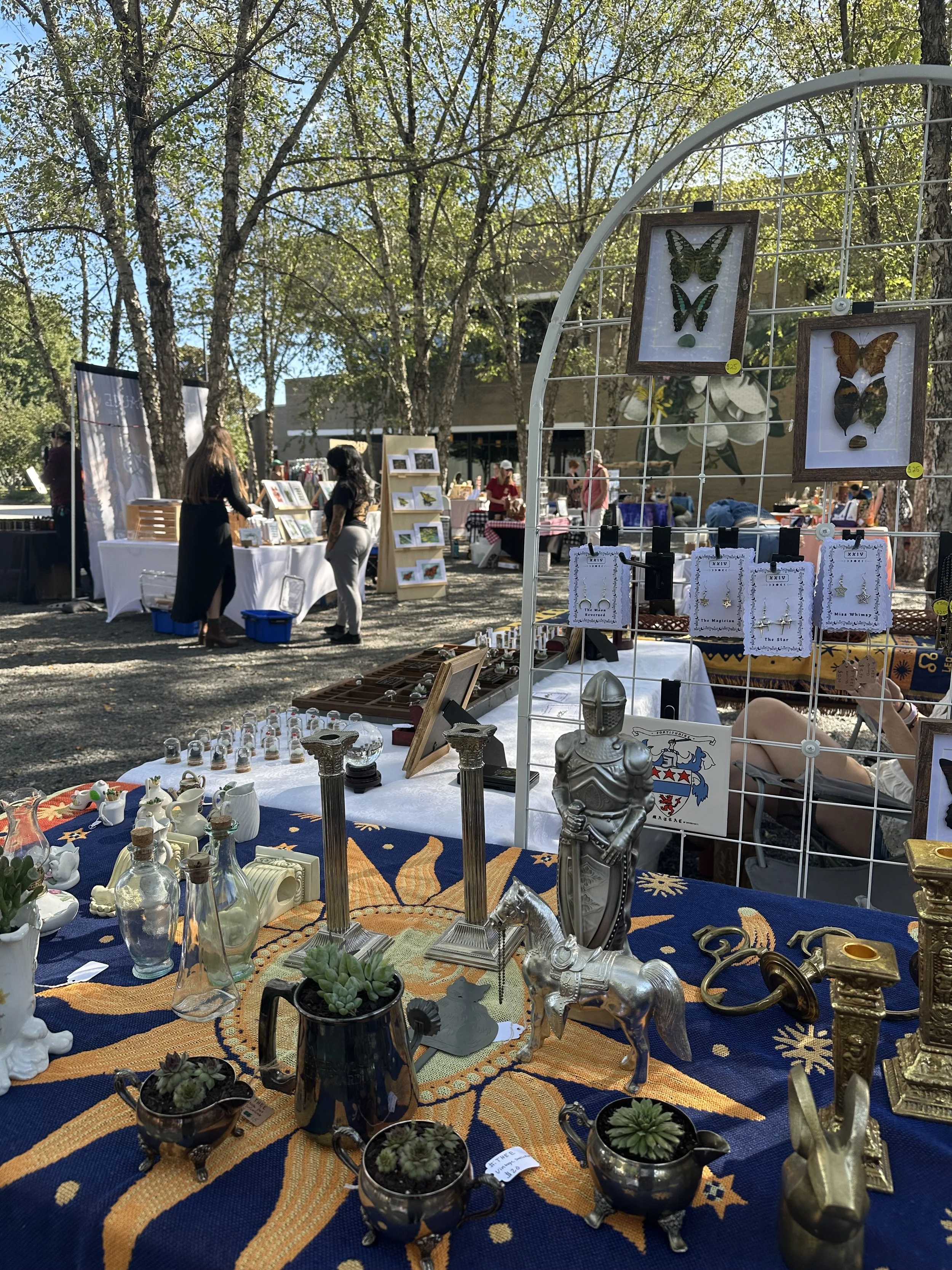 Outdoor market stall selling decorative antiques, jewelry, and framed butterfly art, with people browsing in the background amidst trees and sunlight.