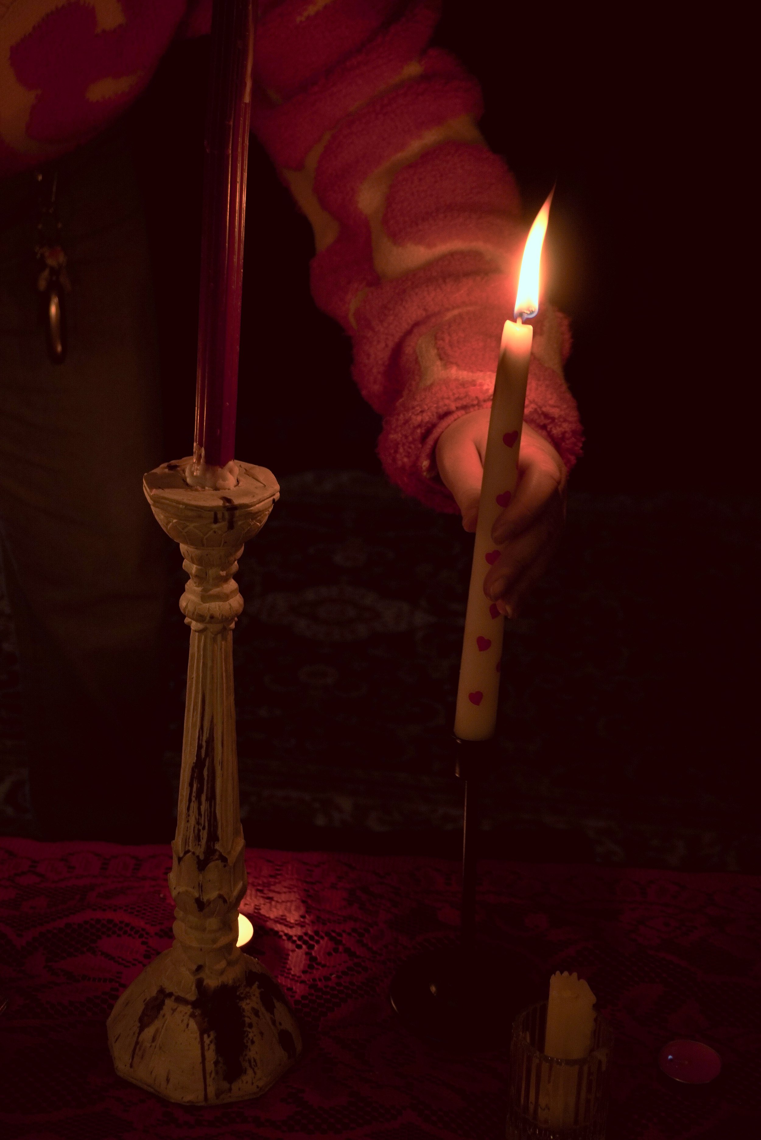 Person holding a lit candle with heart patterns on it in a dark setting, with a tall decorative candle holder and other small candles on a table covered with a lace cloth.
