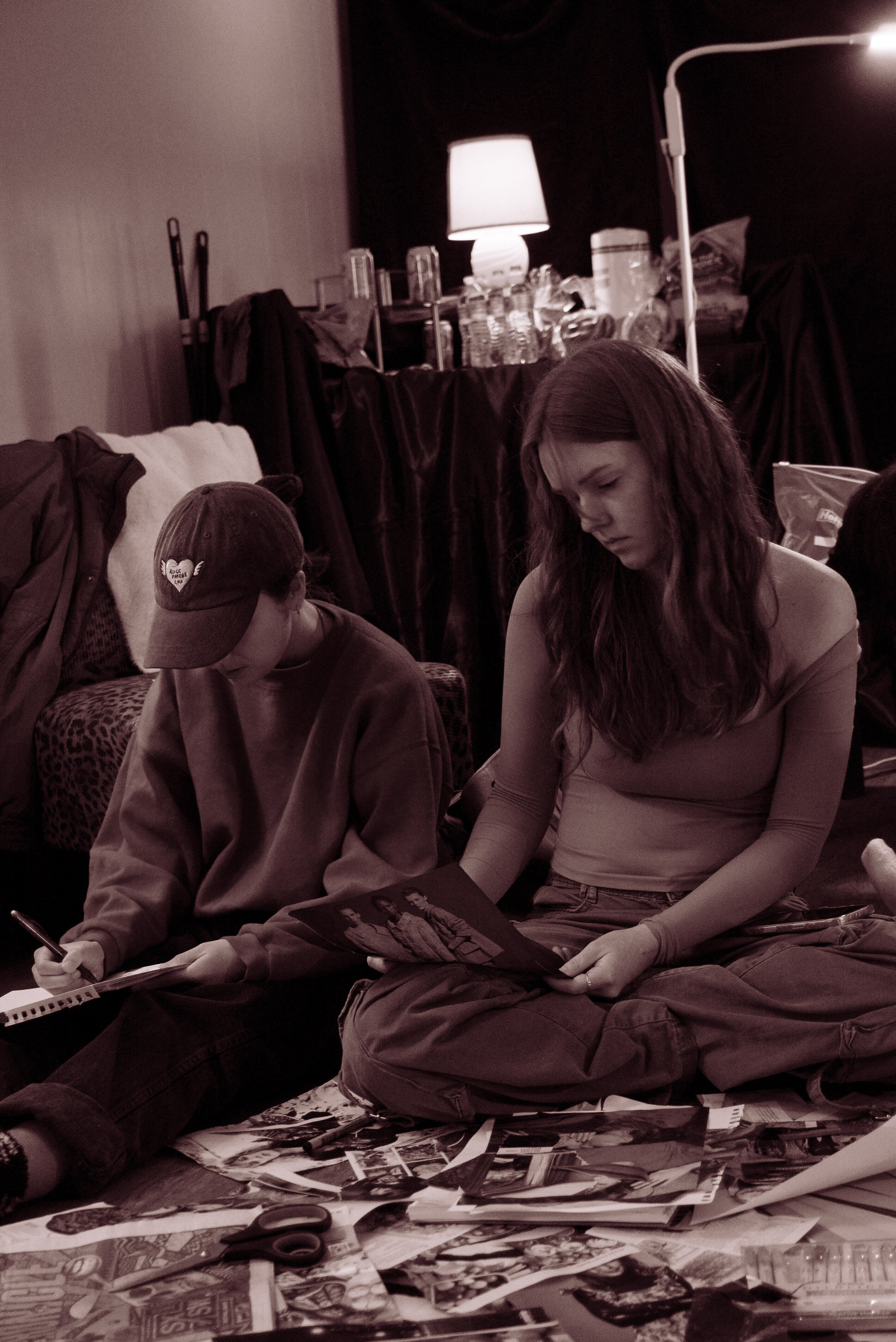 Two people sitting on the floor, looking at photographs. The person on the left is wearing a cap and a sweatshirt, taking notes. The person on the right has long hair and is wearing a long-sleeved top, holding a photograph. The background has a table with water bottles, a lamp, and various items, with clothes draped over furniture.
