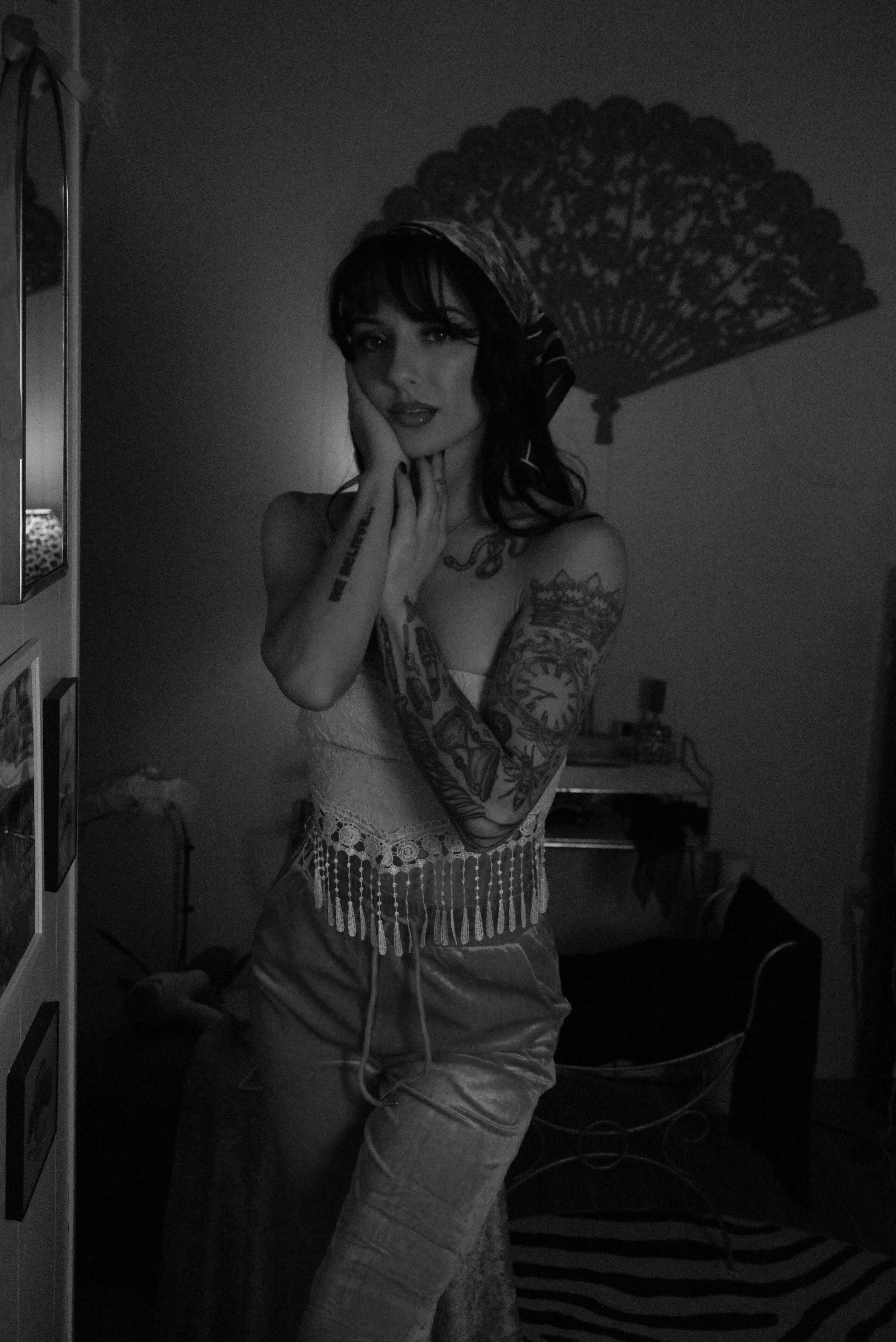 A woman with dark hair and tattoos posing indoors, with a patterned umbrella-shaped shadow on the wall behind her.