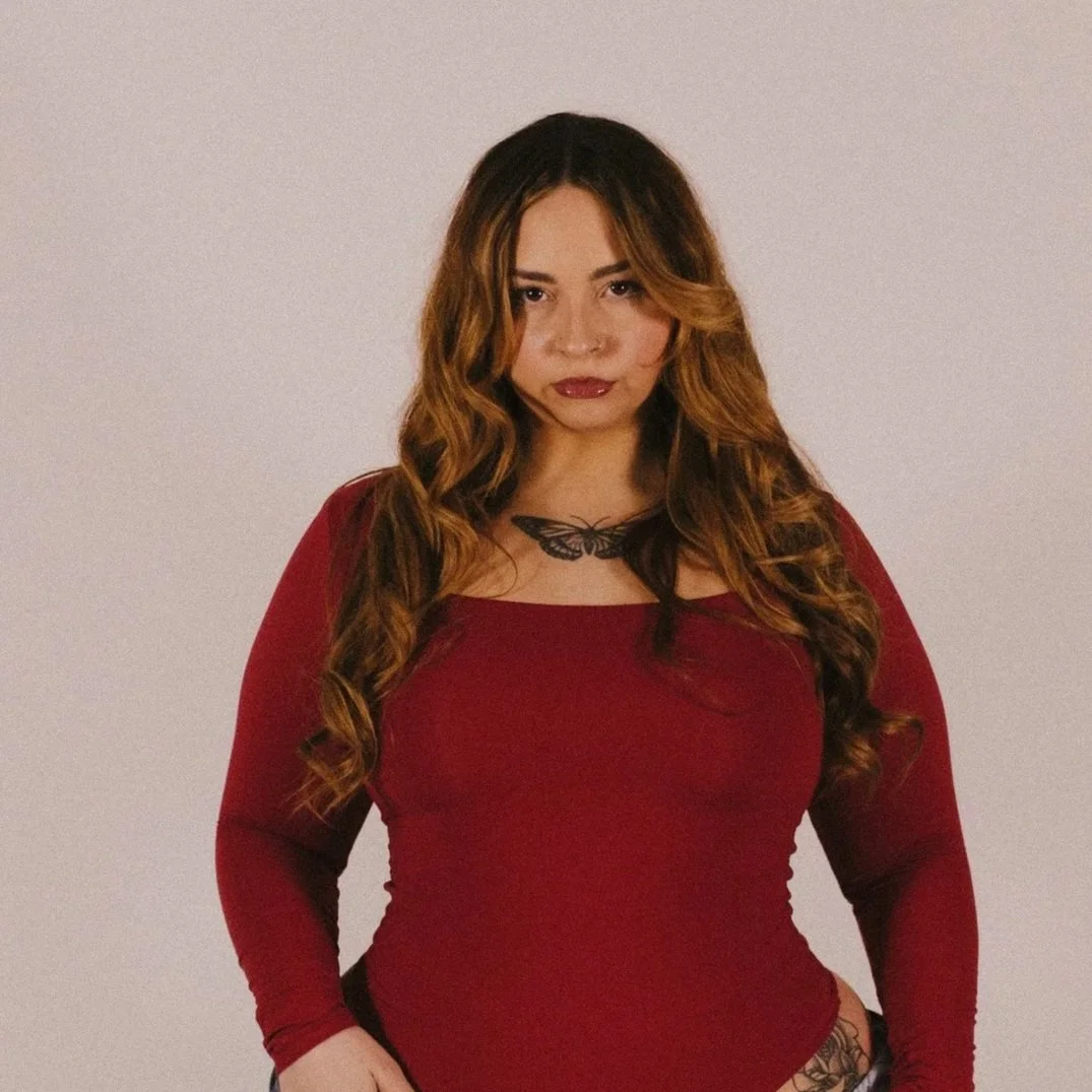 A woman with long wavy hair wearing a red long-sleeve top, standing against a plain background.