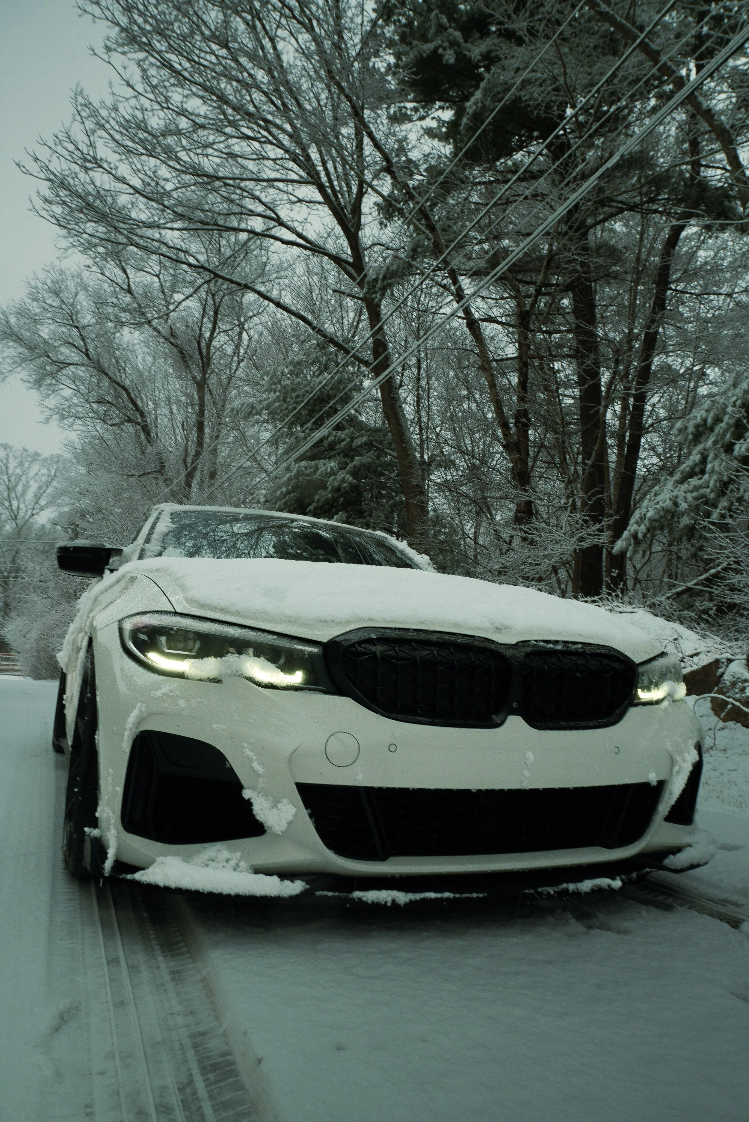 A white car covered in snow parked on a snow-covered street during winter, with snow-laden trees and power lines in the background.