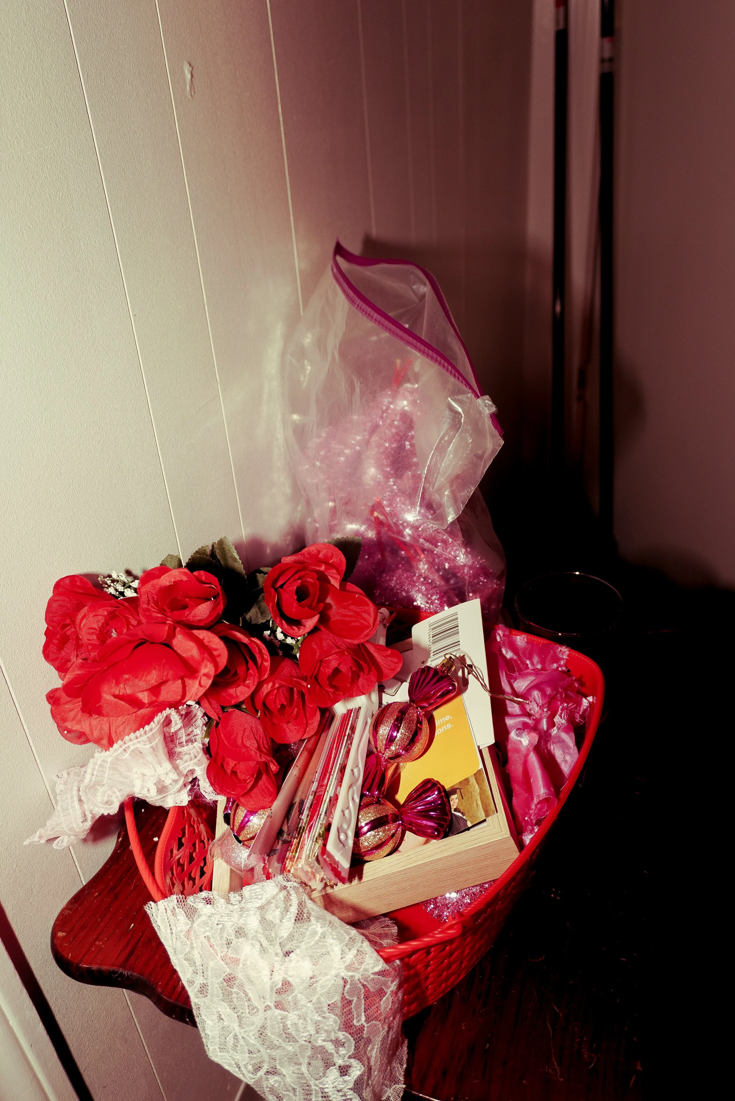A bouquet of red artificial roses, a small box with pink decorative items, and a plastic zippered bag with glitter and pink items inside, arranged on a small wooden table with a lace cloth.
