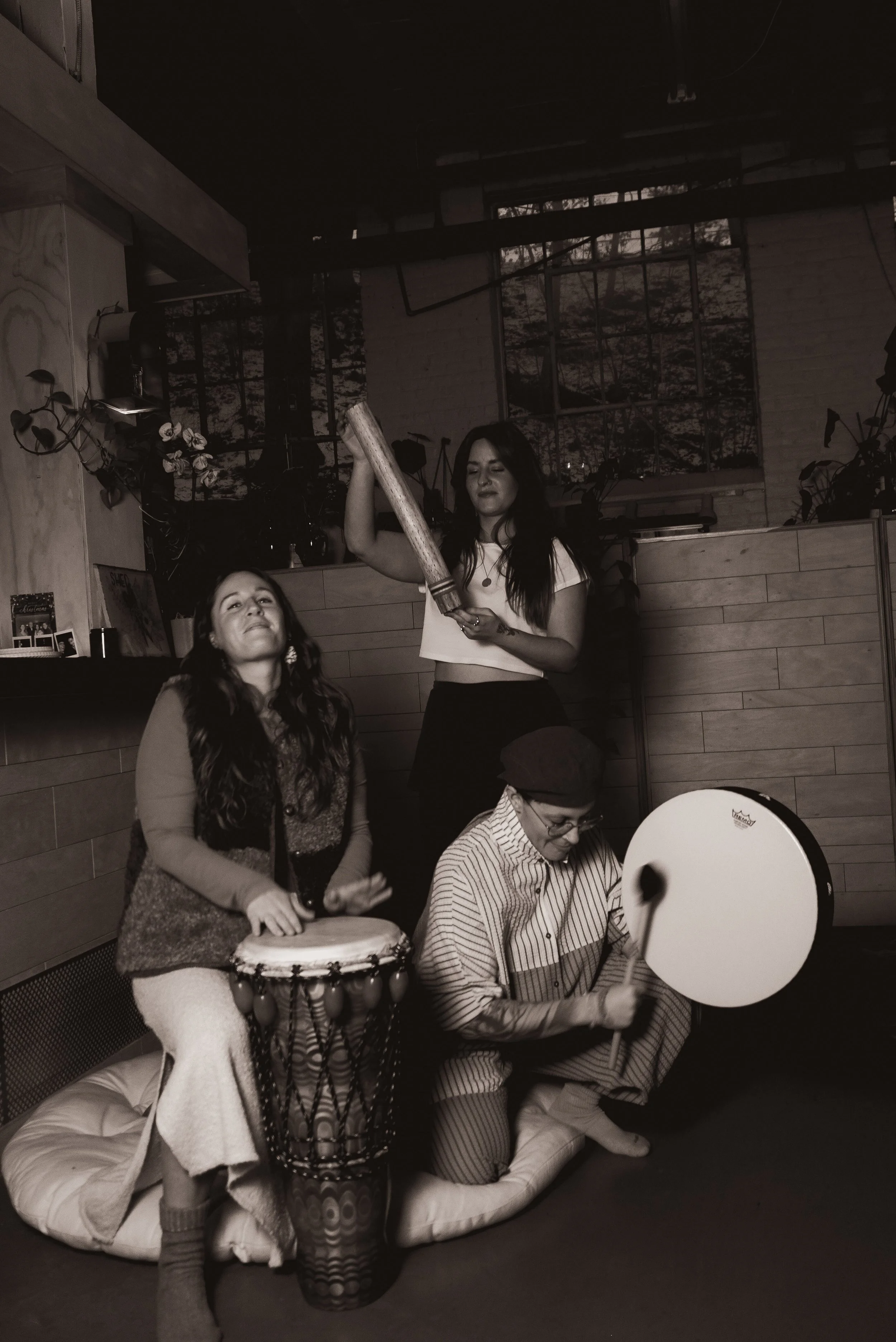 Three people performing musical instruments indoors. Two women, one on a drum and the other holding a large stick or mallet, and a man playing a large tambourine or frame drum. The scene appears lively and artistic, with a cozy setting and plants in the background.