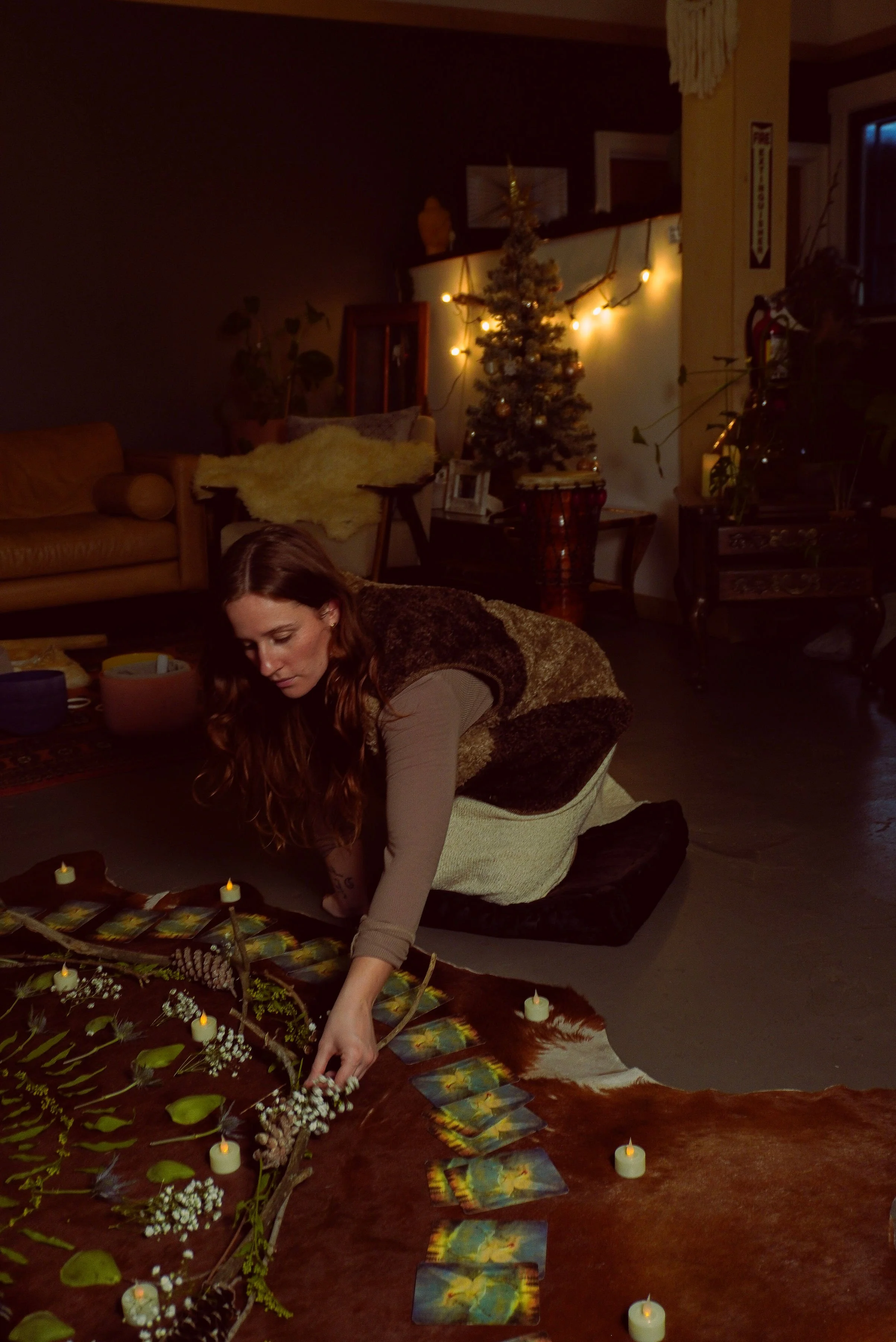 A woman with long red hair arranging tarot cards in a dimly lit room. The background features a decorated Christmas tree with lights, a yellow couch with a fuzzy blanket, and various plants and furniture.