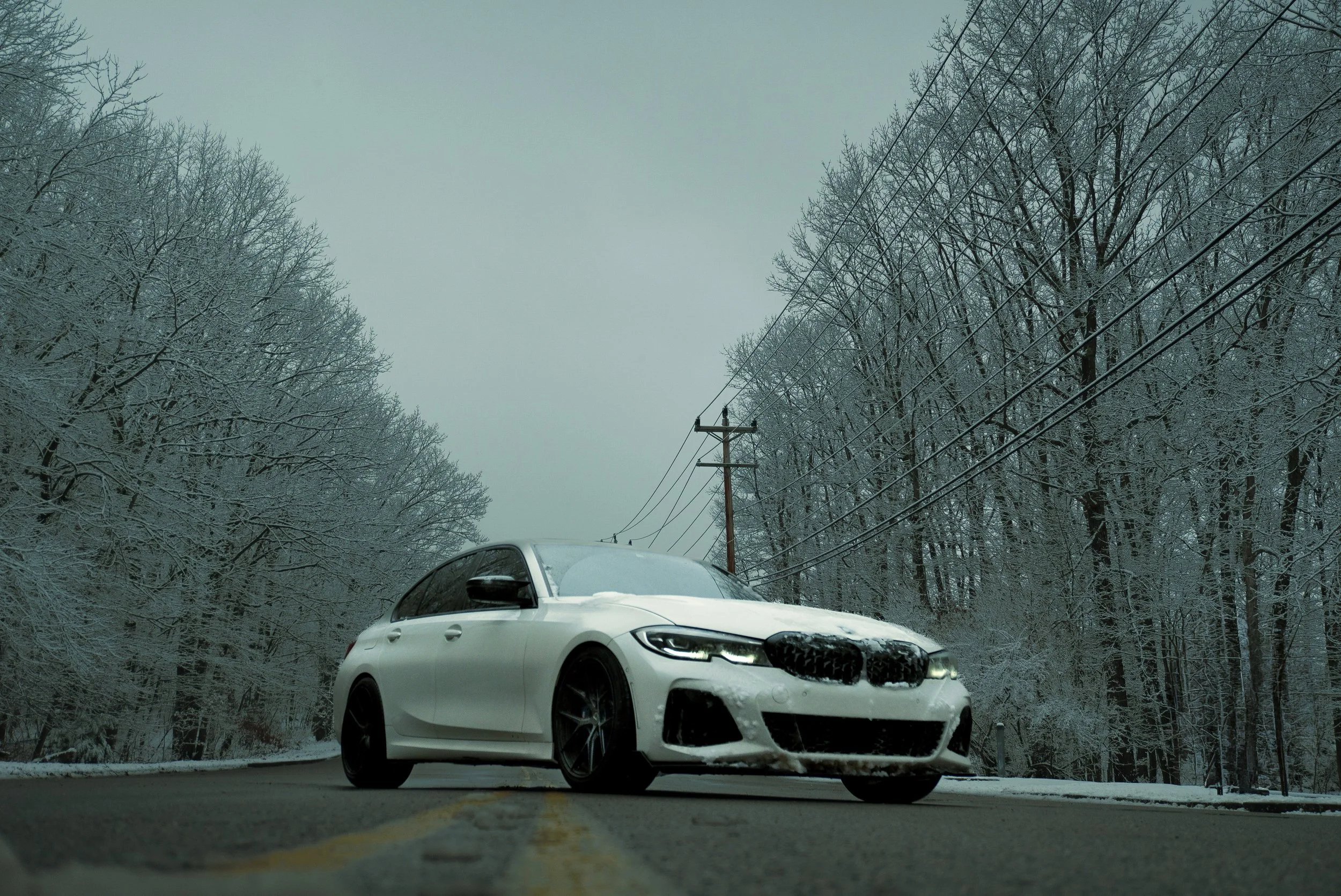 White BMW sedan parked on a snow-covered road surrounded by snow-dusted trees and a utility pole with power lines, on a cloudy winter day.