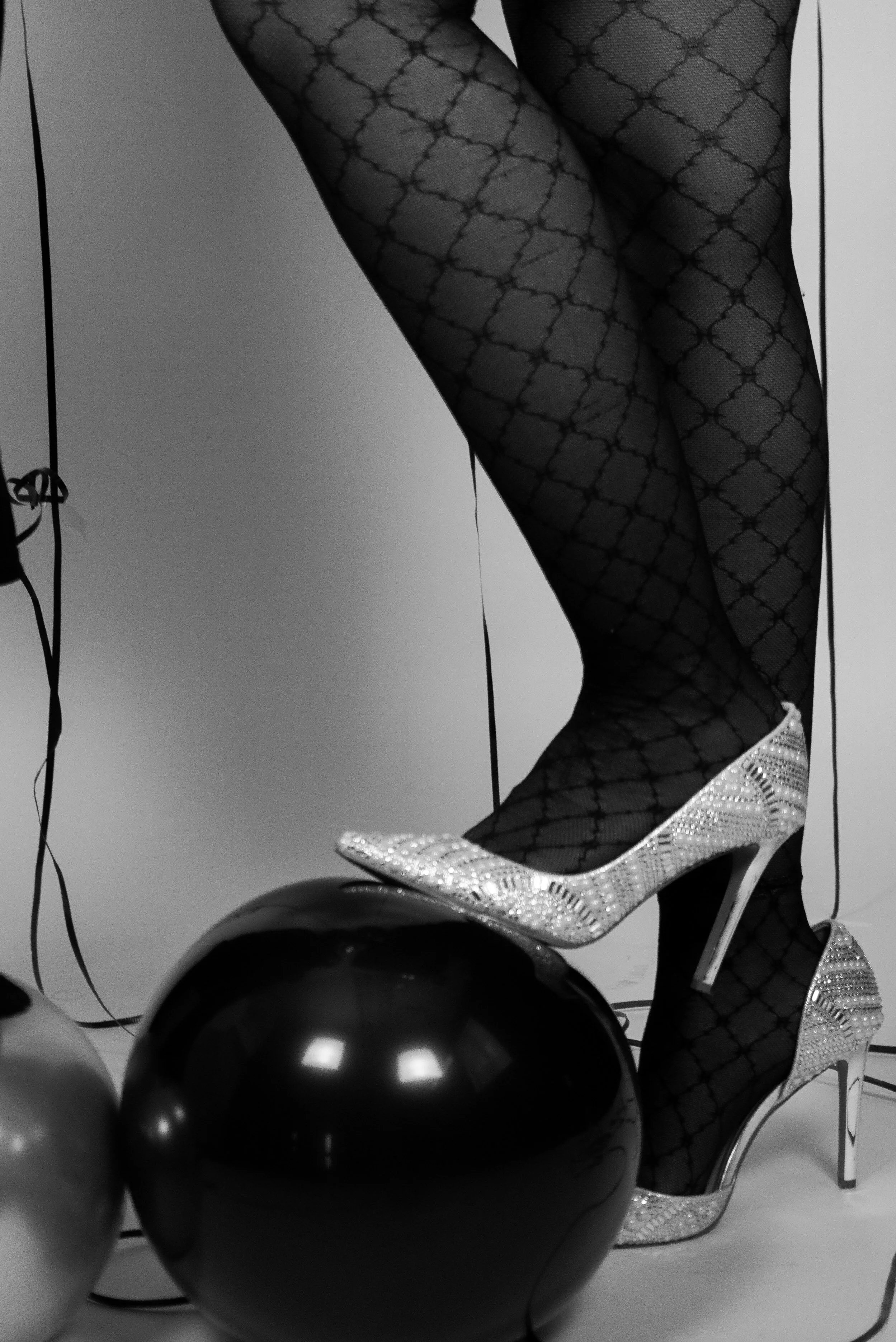 Close-up of a woman's legs wearing patterned fishnet stockings and high-heeled shoes with rhinestones, with balloons in the foreground.