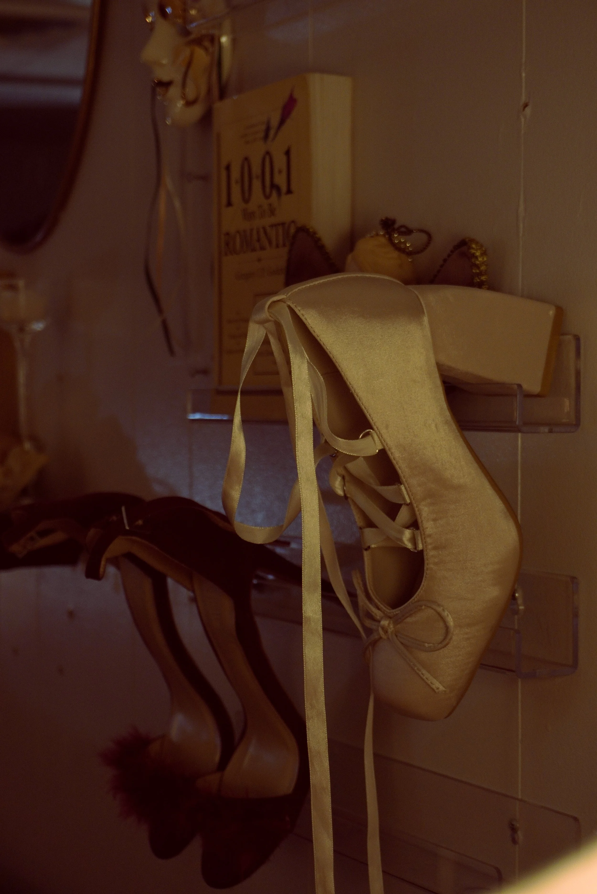 A pair of satin ballet pointe shoes hanging from a clear plastic holder on a wall, with other mounted items and a mirror in the background.