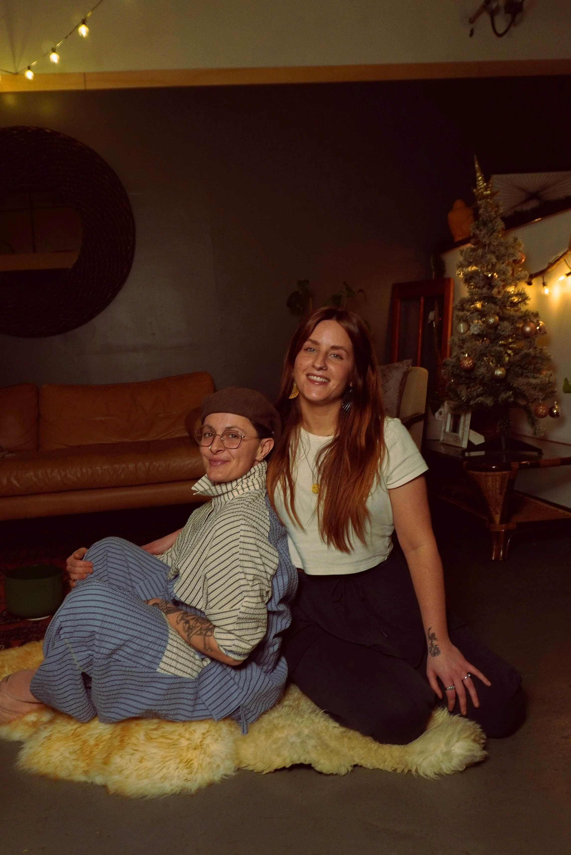 Two women sitting on a fluffy rug in a cozy living room decorated with Christmas tree, string lights, and warm lighting, smiling at the camera.
