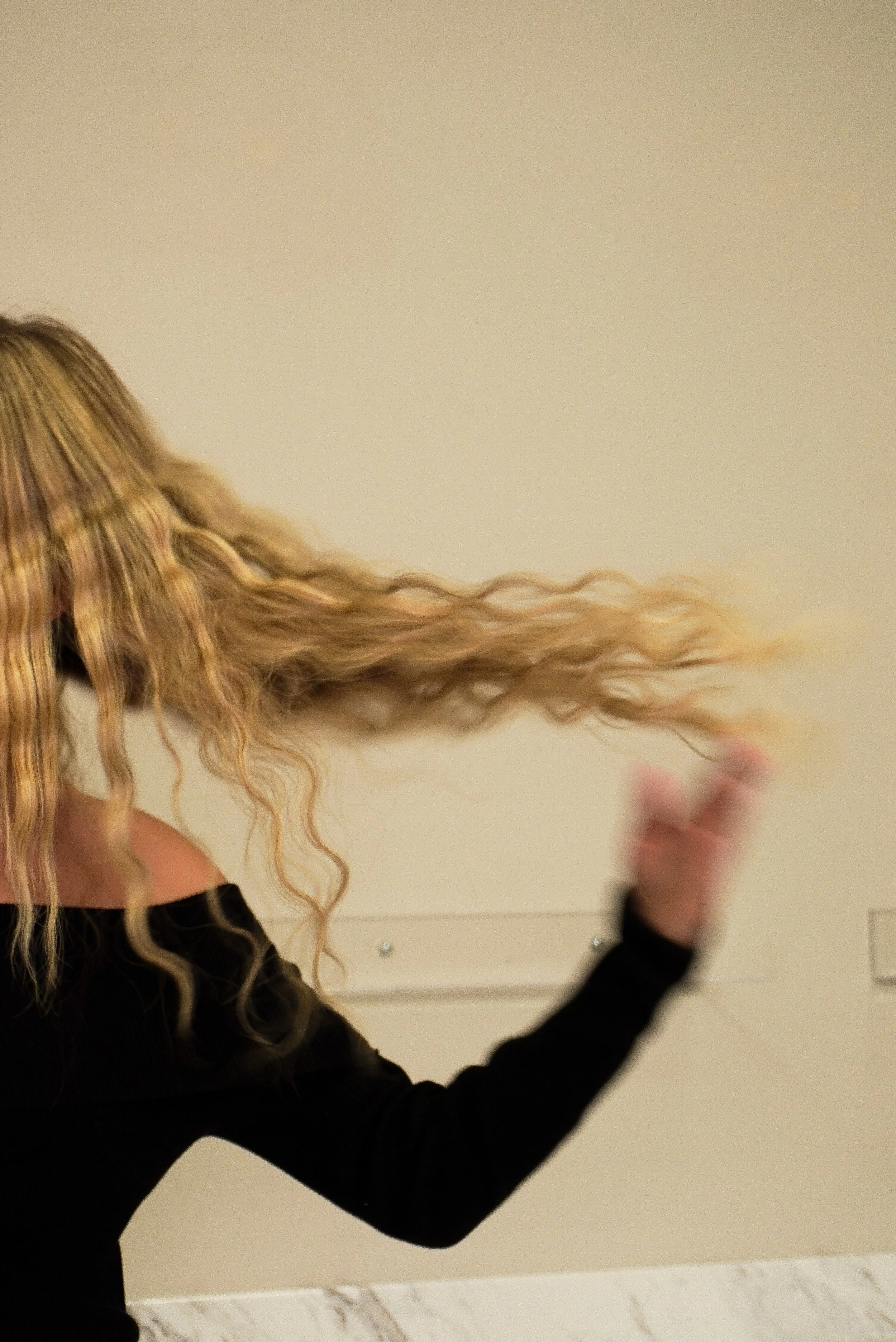 A person with long, wavy blonde hair flips their hair, wearing a black long-sleeve top, against a light-colored wall.