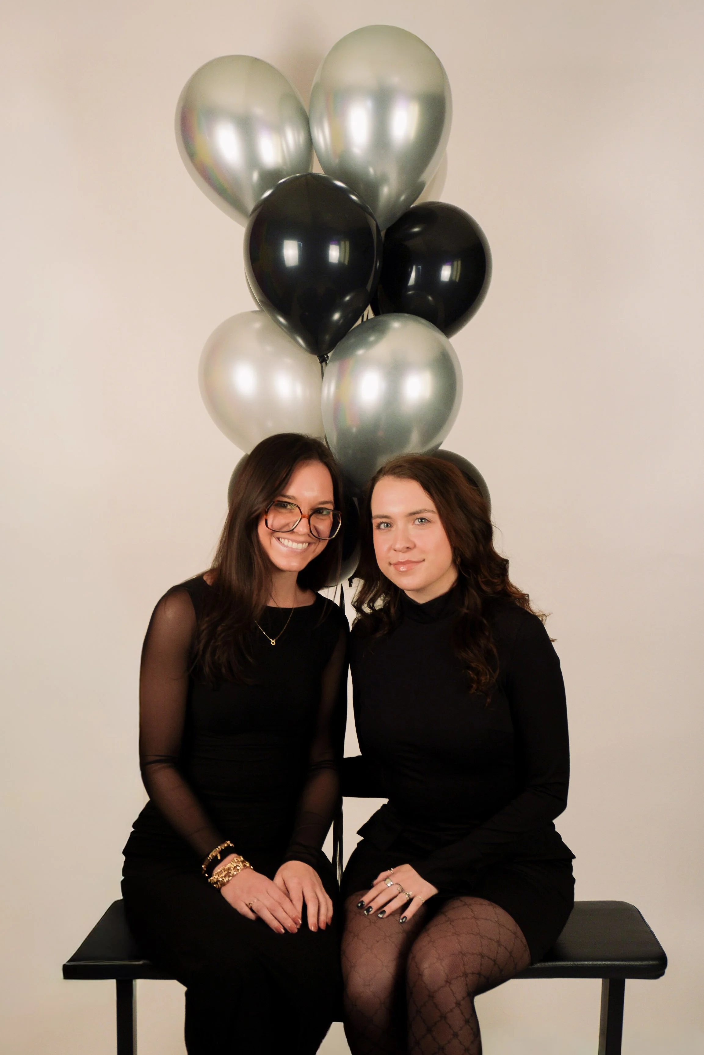 Two women sitting on a black bench in front of a white wall, with a bunch of black, silver, and gray balloons behind them.
