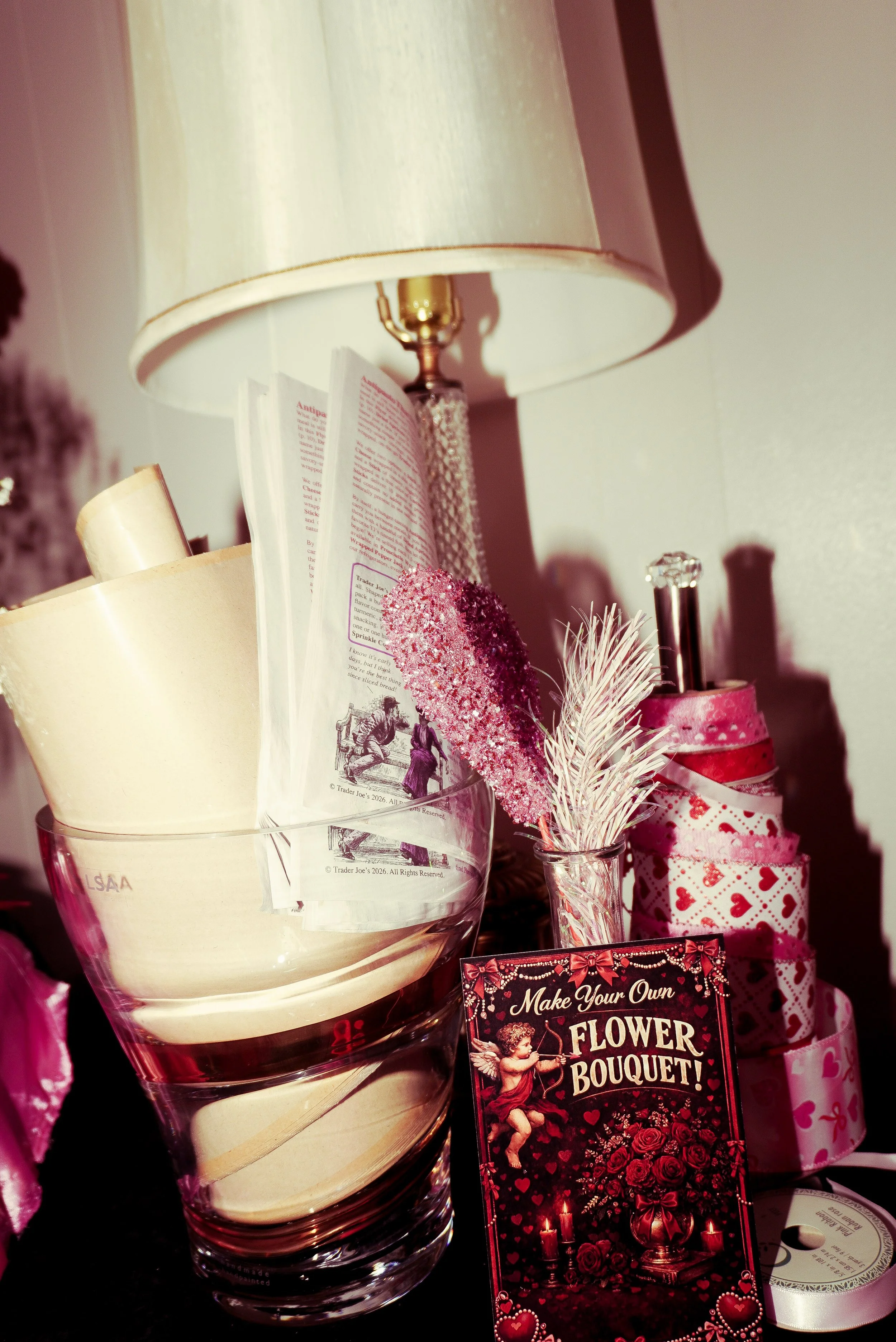 A pink-themed decorative display with a lamp, a glass bowl holding paper and a cream-colored object, a small vase with pink and white synthetic flowers, a card titled "Make Your Own Flower Bouquet!", and a pink gift-wrapping roll with heart patterns.