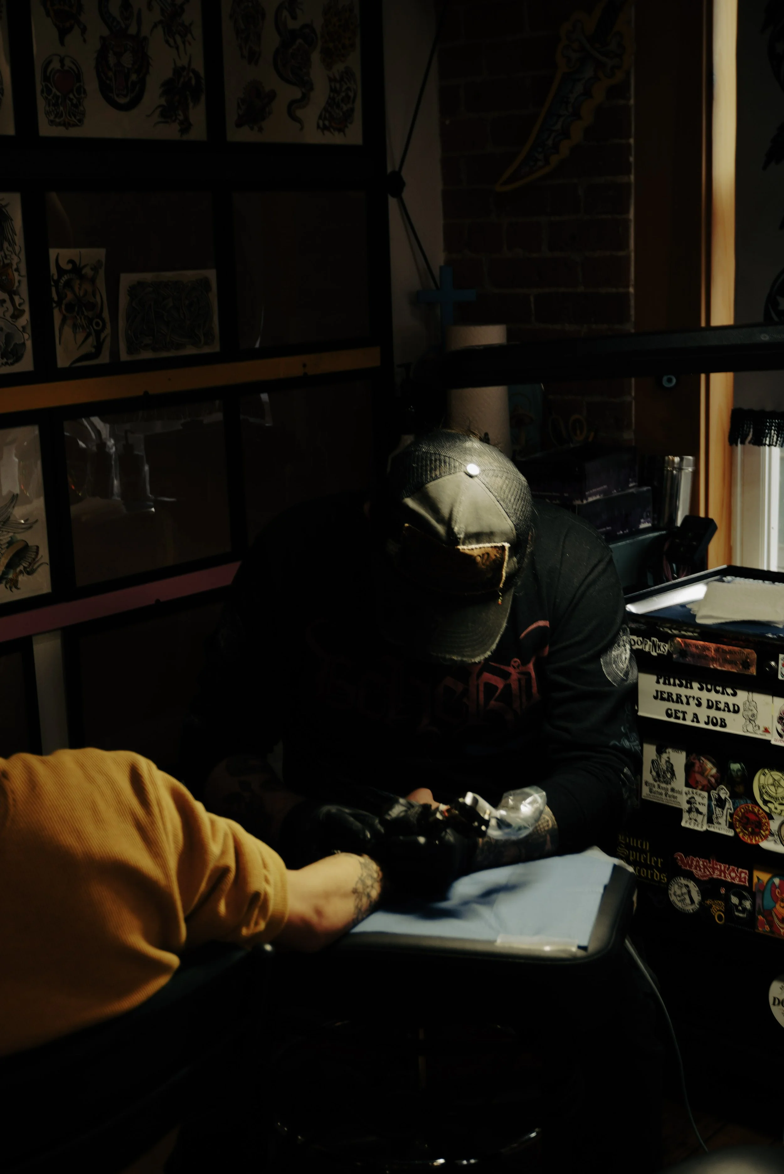A person with a tattooed arm and black gloves appears to be tattooing another person's arm in a dimly lit room decorated with tattoo stickers and graffiti art.