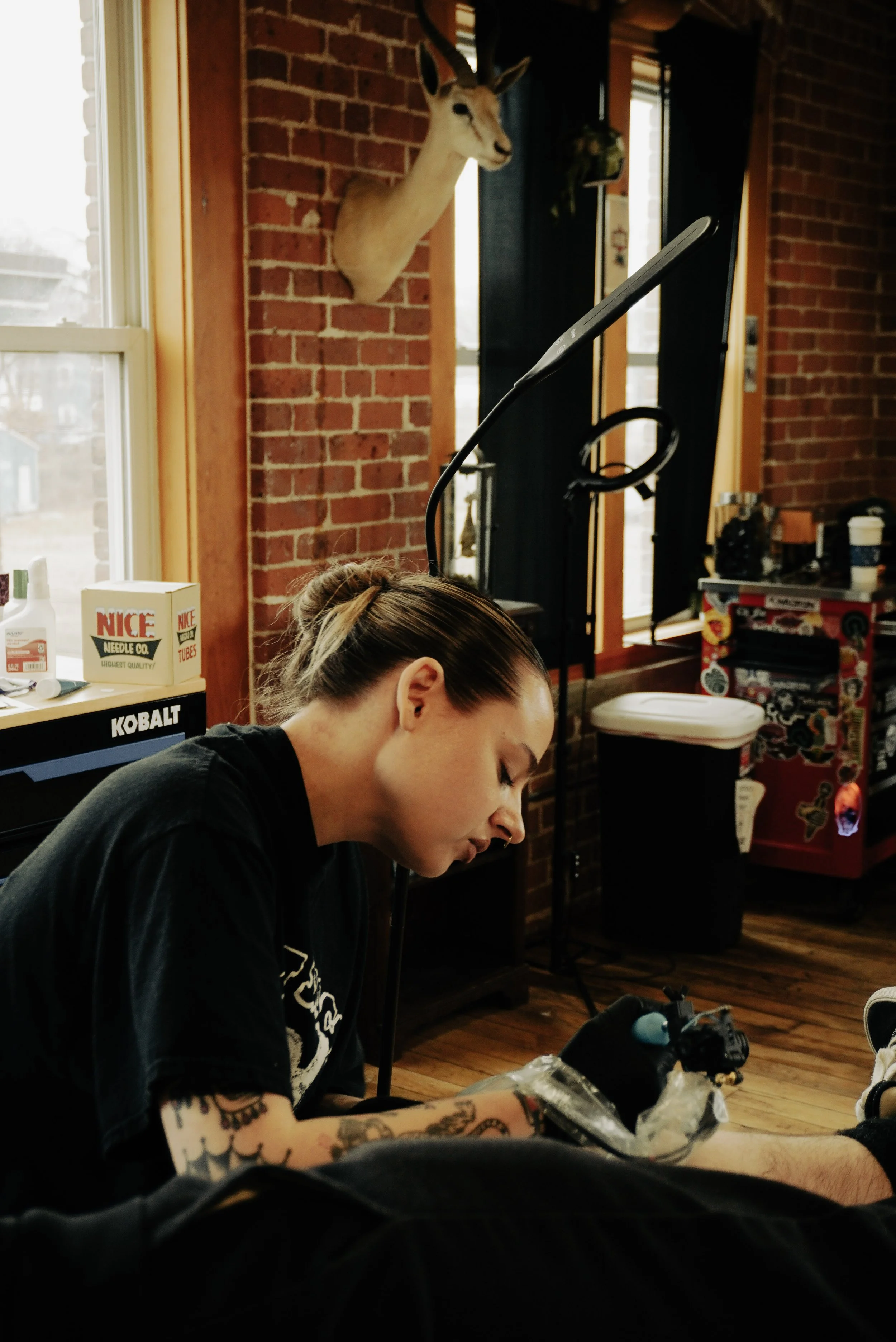 A woman with tattoos doing a tattoo on someone's leg in a tattoo shop, with a deer head mounted on a brick wall behind her.