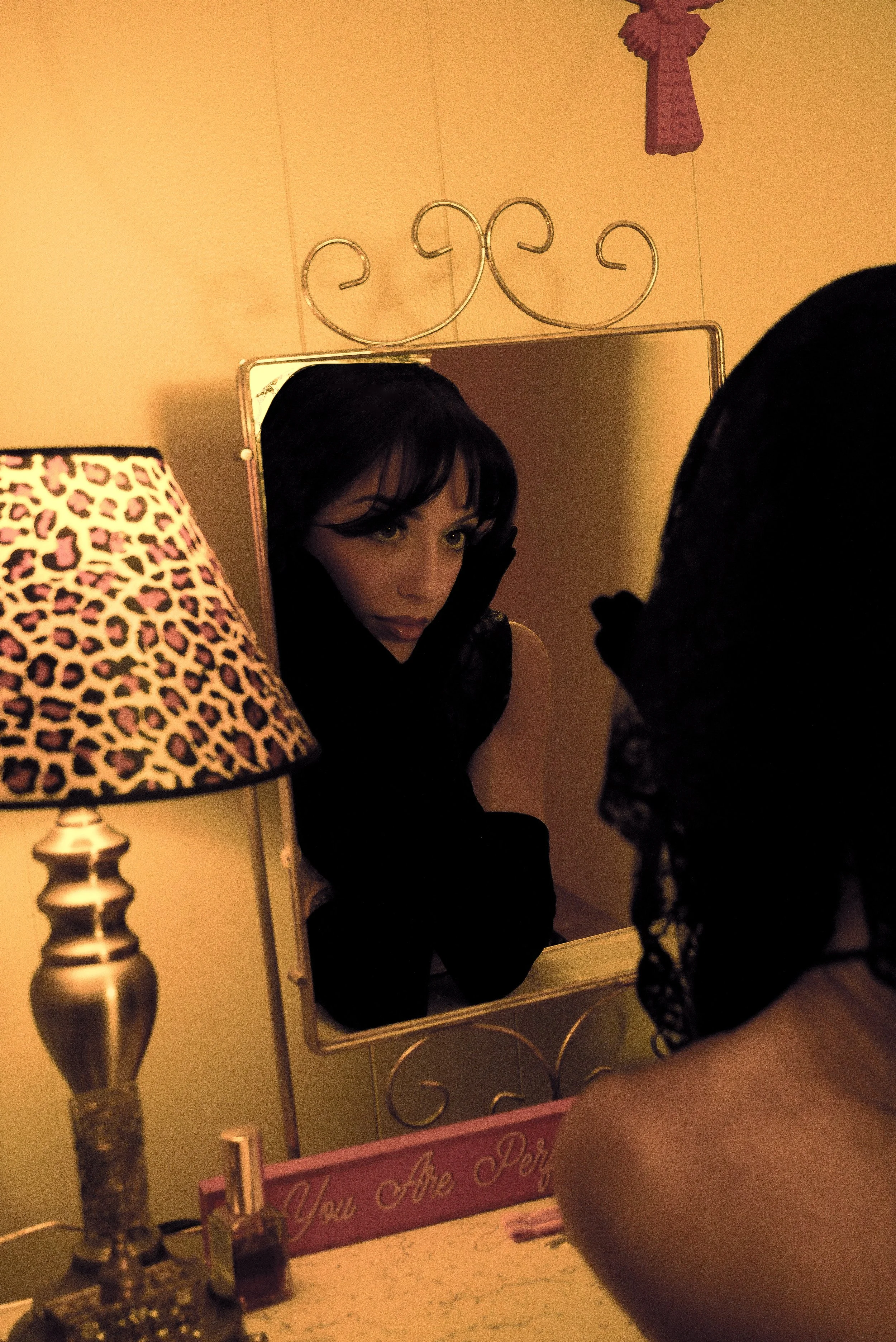 A woman looking into a mirror, resting her face on her hand, with a leopard-print lamp to the side and a pink sign on the table that says 'You Are Perfect'.