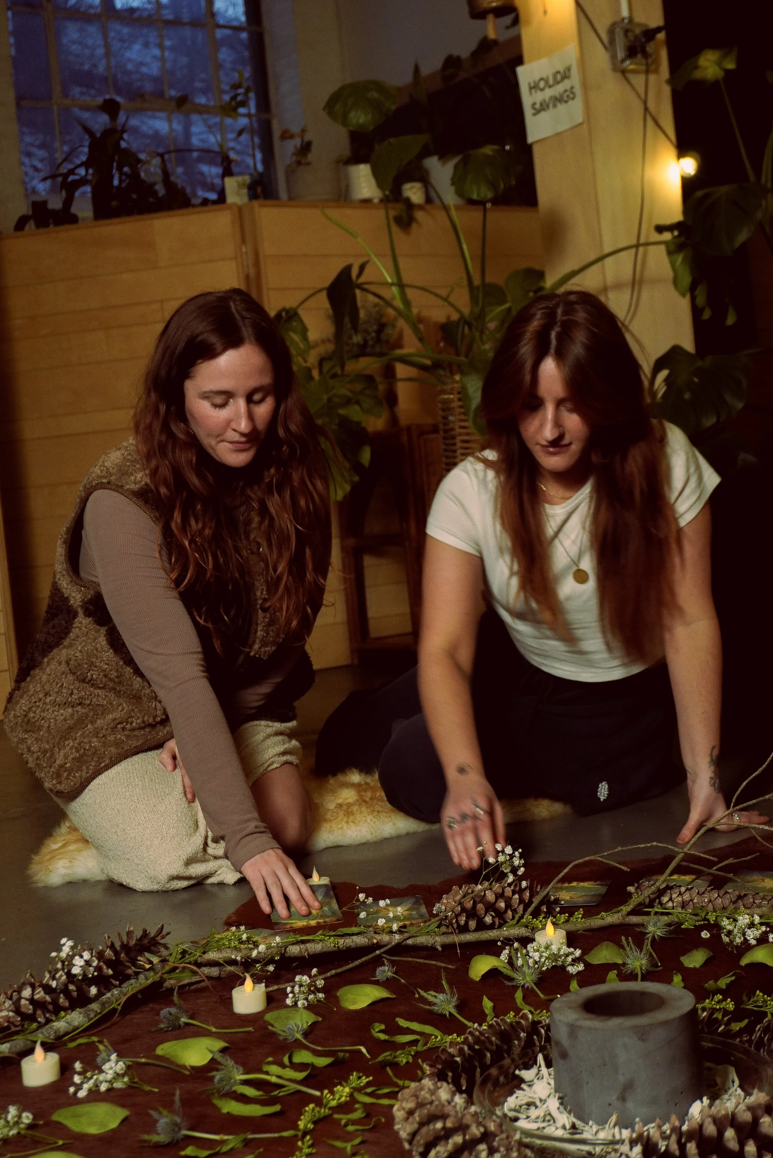 Two women sit on the floor amid candles, pinecones, leaves, and branches, creating a nature-inspired arrangement in a cozy indoor space with large plants and warm lighting.